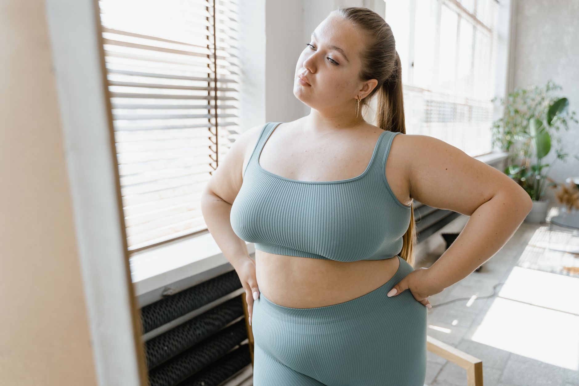 A plus-size woman in activewear poses confidently indoors with hands on her waist.