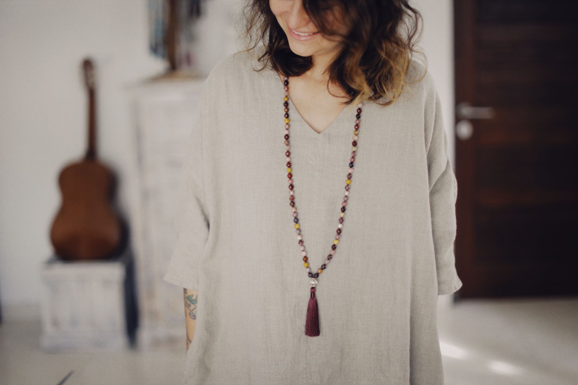 A woman stands indoors wearing a long multicolored beaded necklace, expressing casual style and elegance.