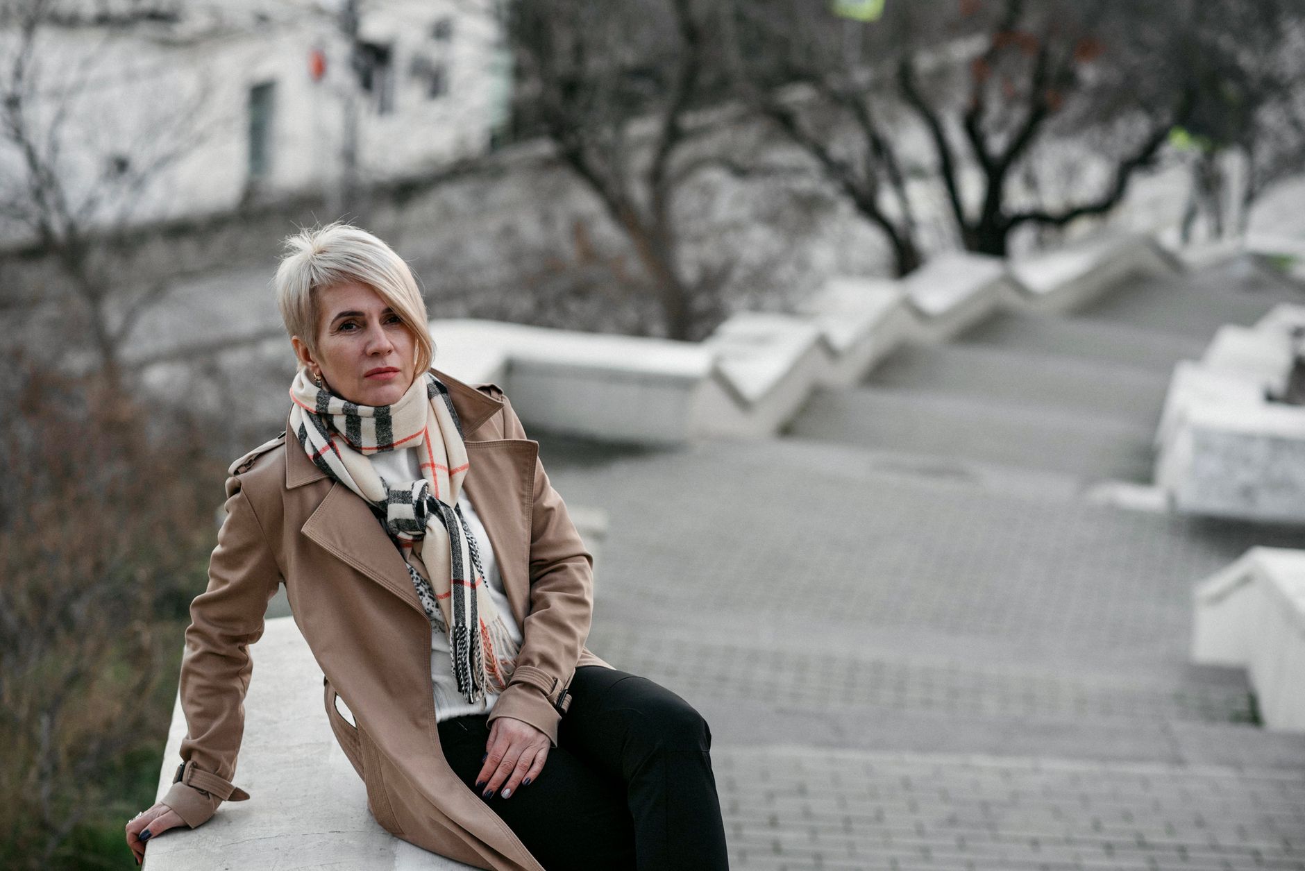Blonde woman in a trench coat posing on an outdoor staircase, showcasing urban fashion.