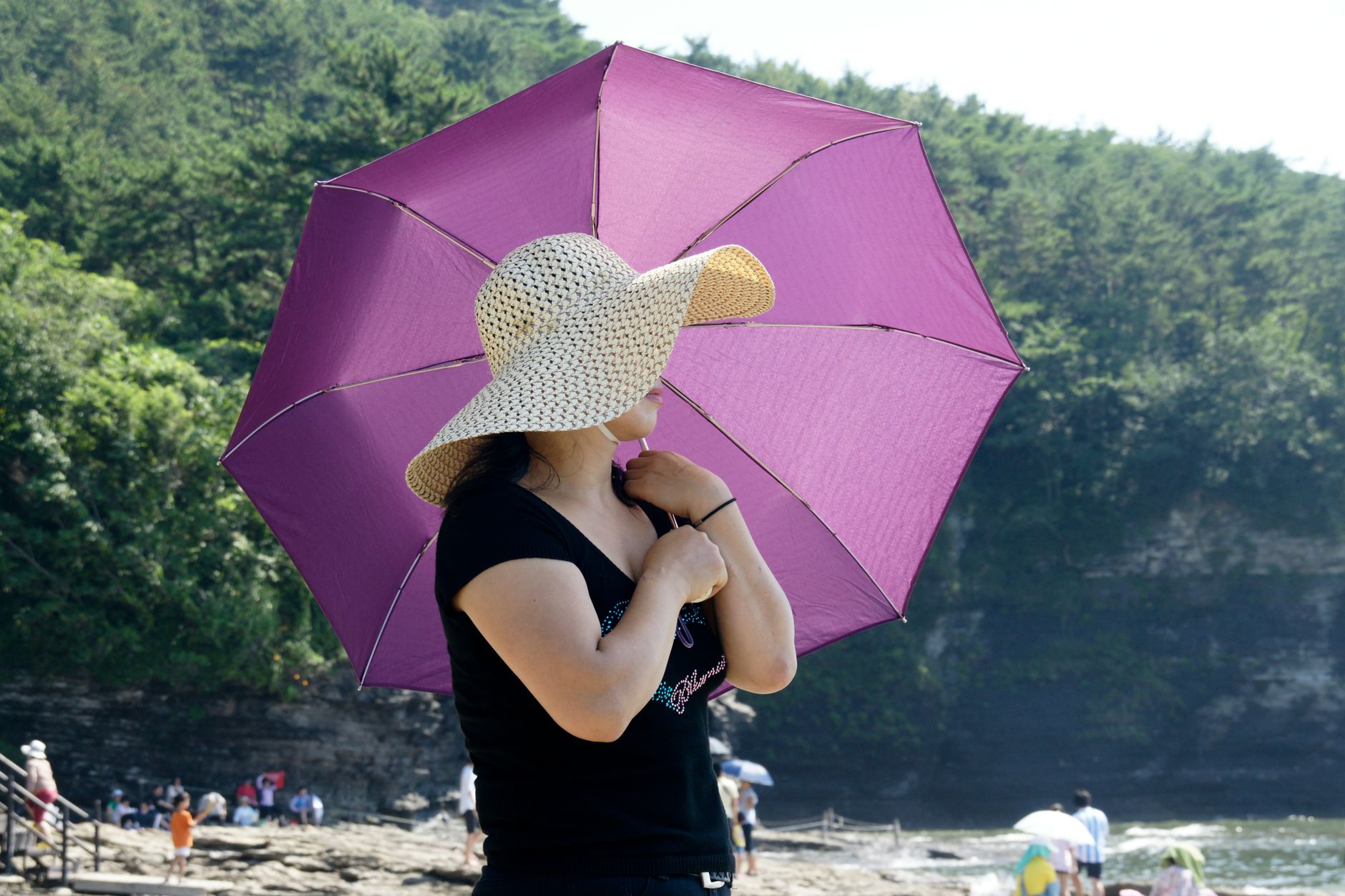 A woman wearing a sun hat holding a pink umbrella on a sunny beach day.