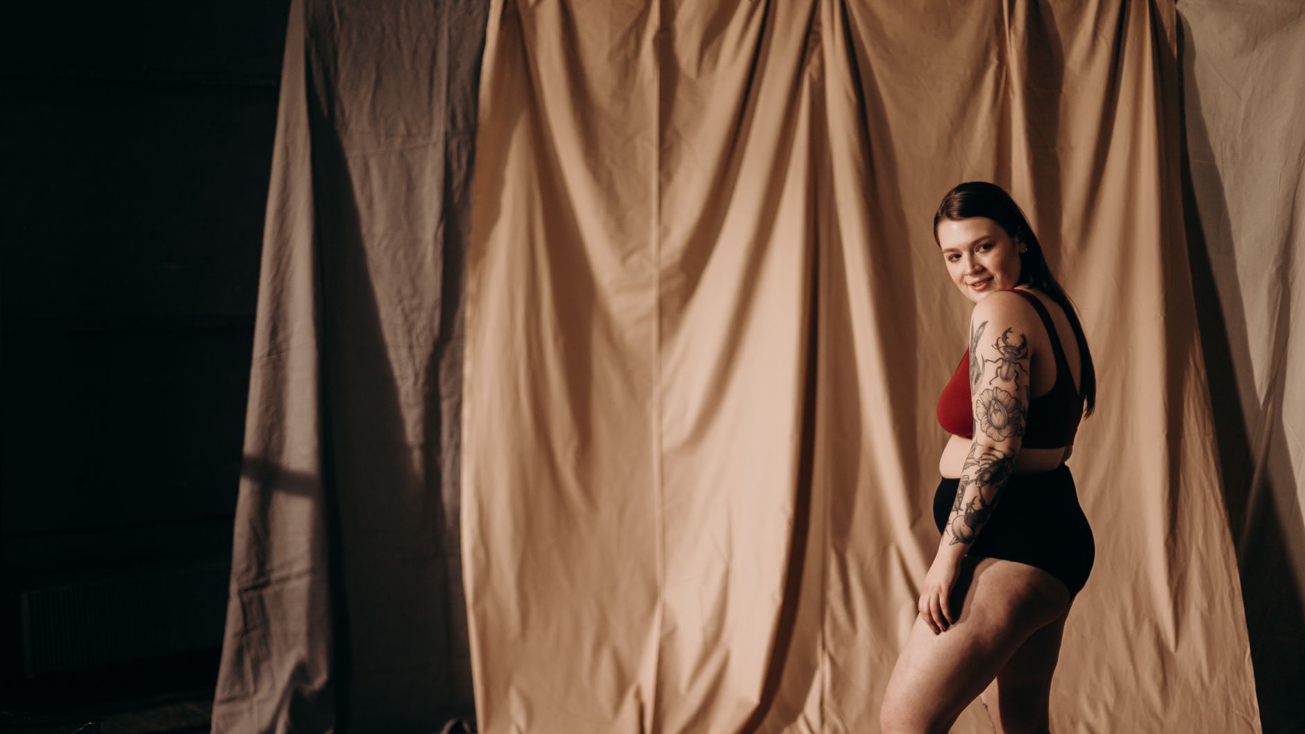 Confident plus-size woman with arm tattoo posing in a studio setting, smiling.