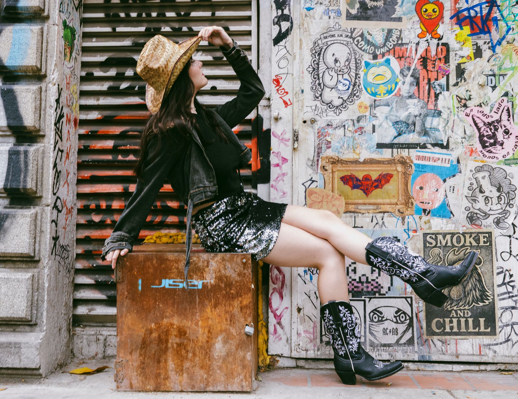 Young woman in cowboy hat and boots poses stylishly against a vibrant graffiti wall.