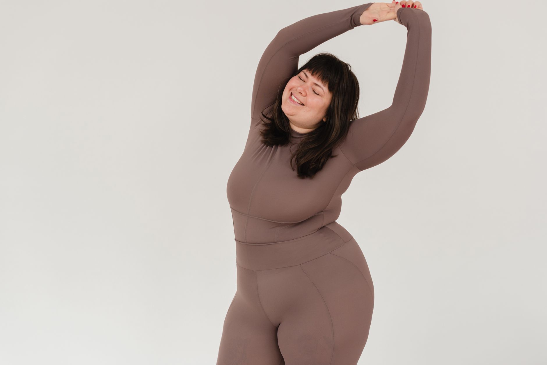 Positive plus size female in sportswear stretching oneself with closed eyes against white background