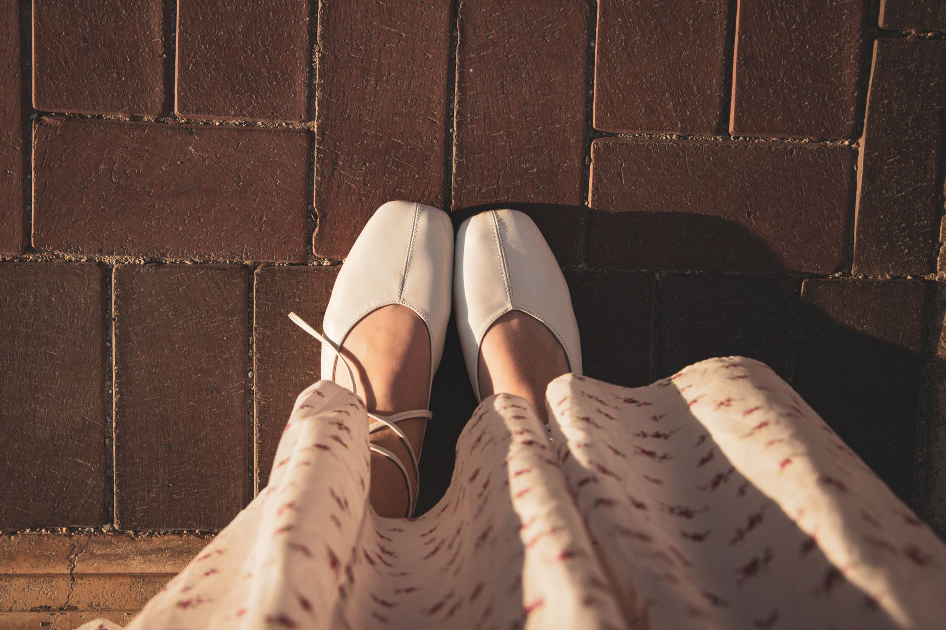 Top-down view of white ballet flats paired with a flowing, patterned dress on a brick pathway.
