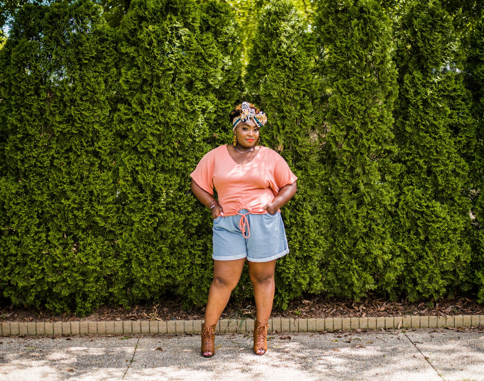 Confident woman posing in summer fashion against a vibrant green hedge.