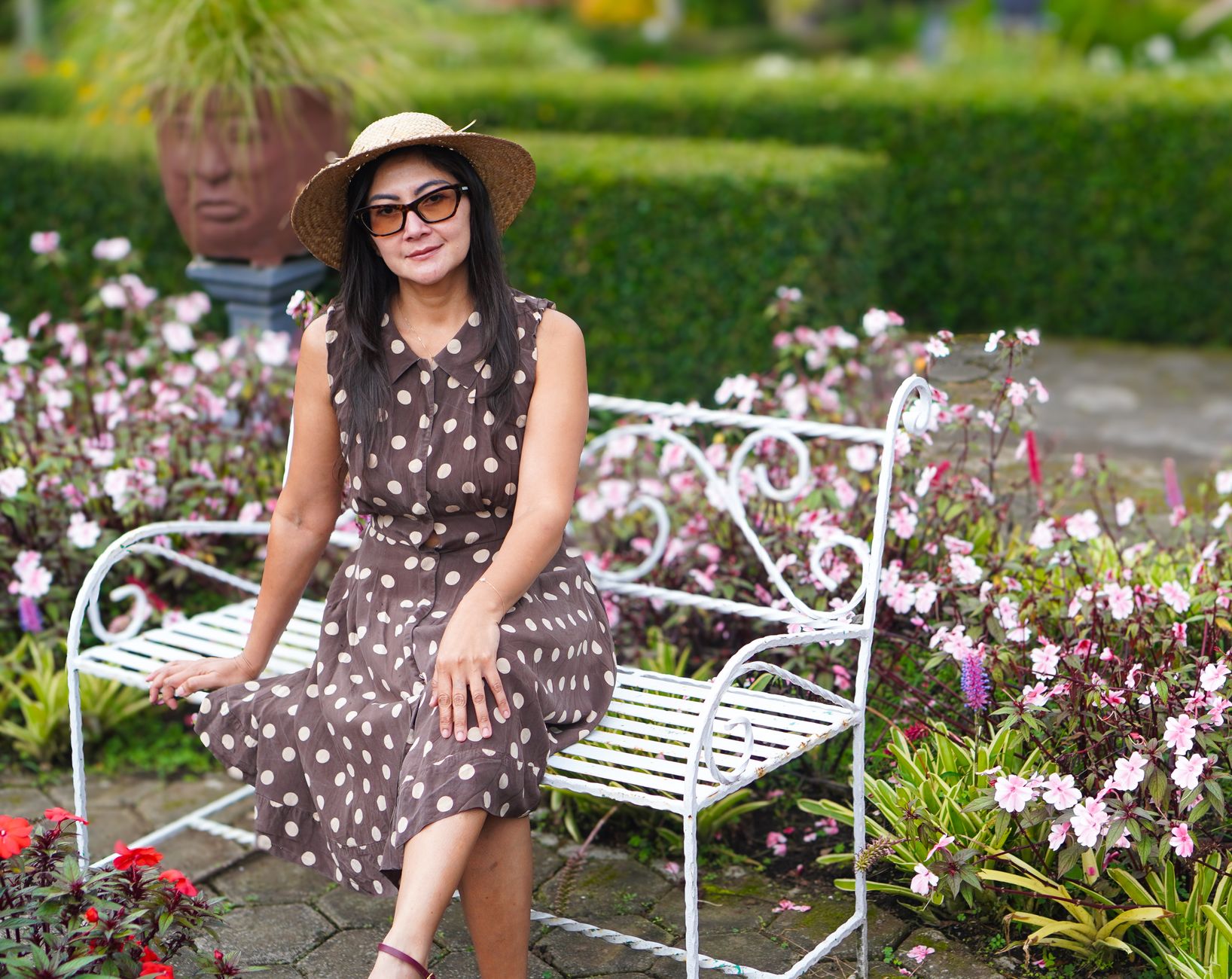 A stylish woman in a polka dot dress and hat sits on a bench surrounded by flowers in a garden.