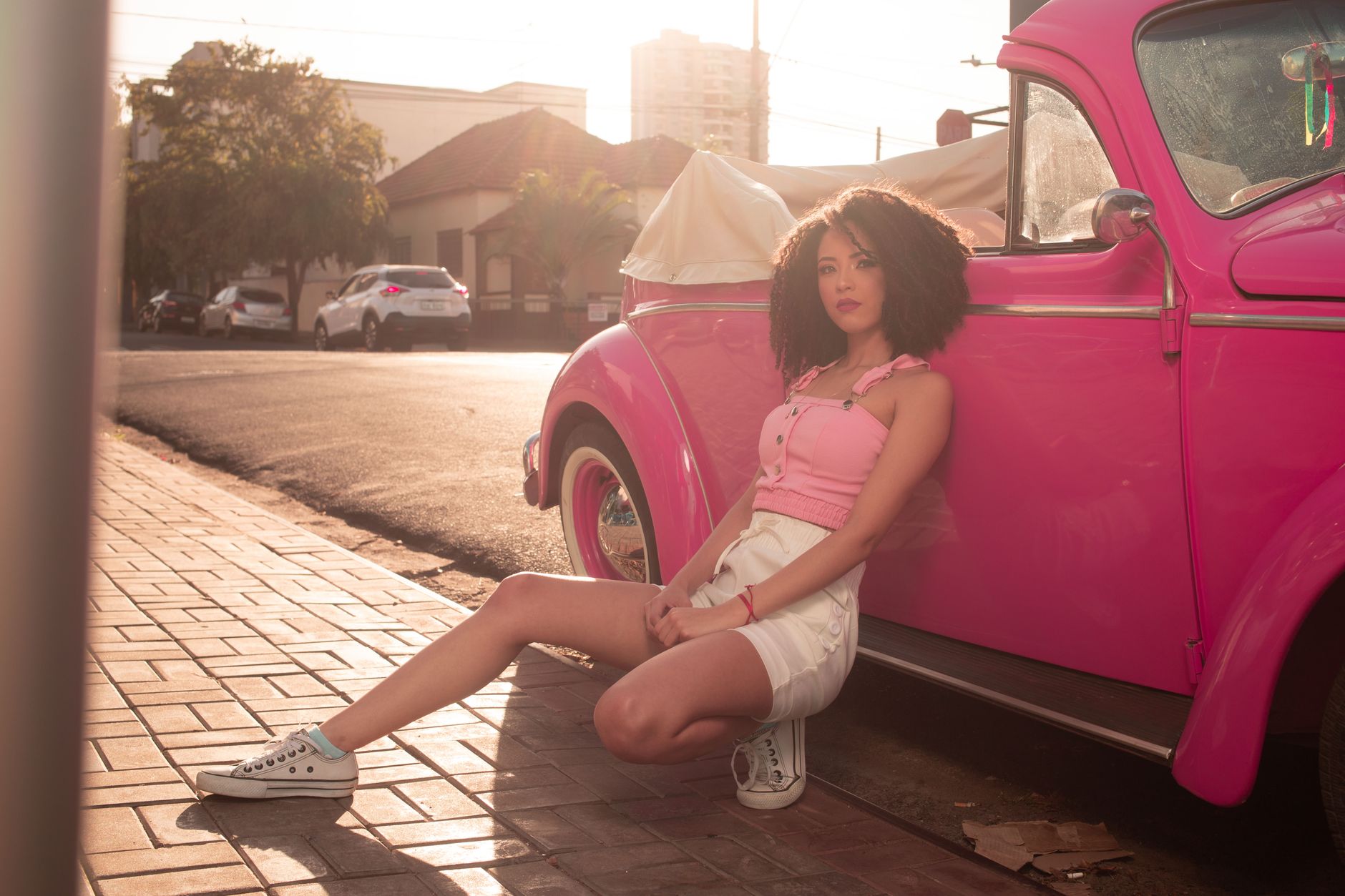 A woman in casual summer attire posing with a pink vintage car on a city street.