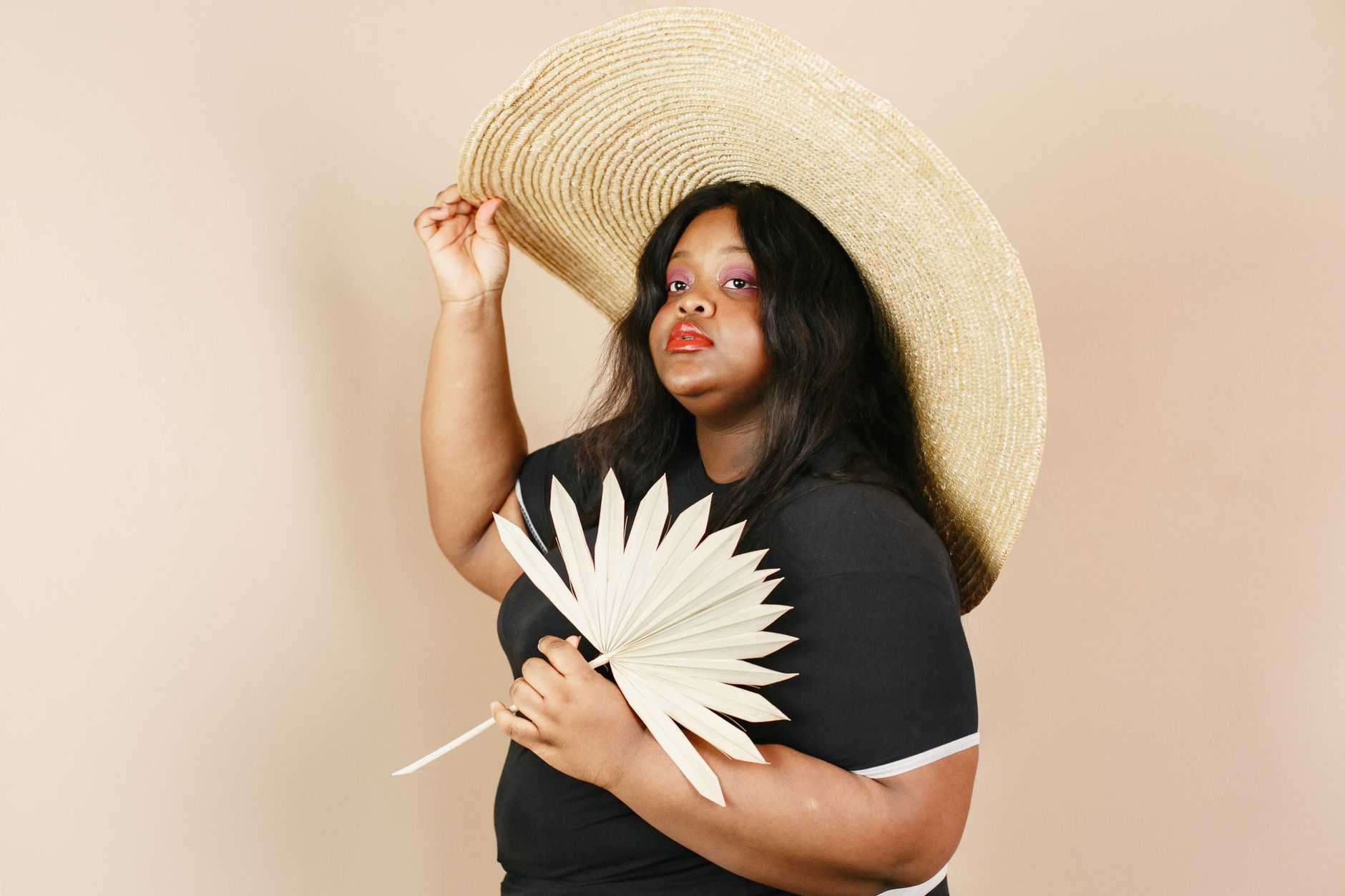 Portrait of a stylish woman holding a dry palm leaf, wearing a large straw hat in a studio setting.