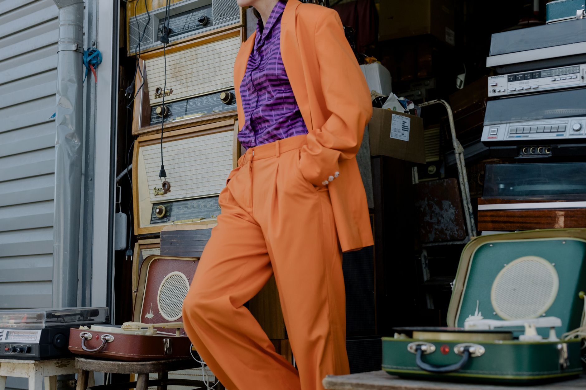 A fashionable person in an orange suit leaning against retro stereos in a store.
