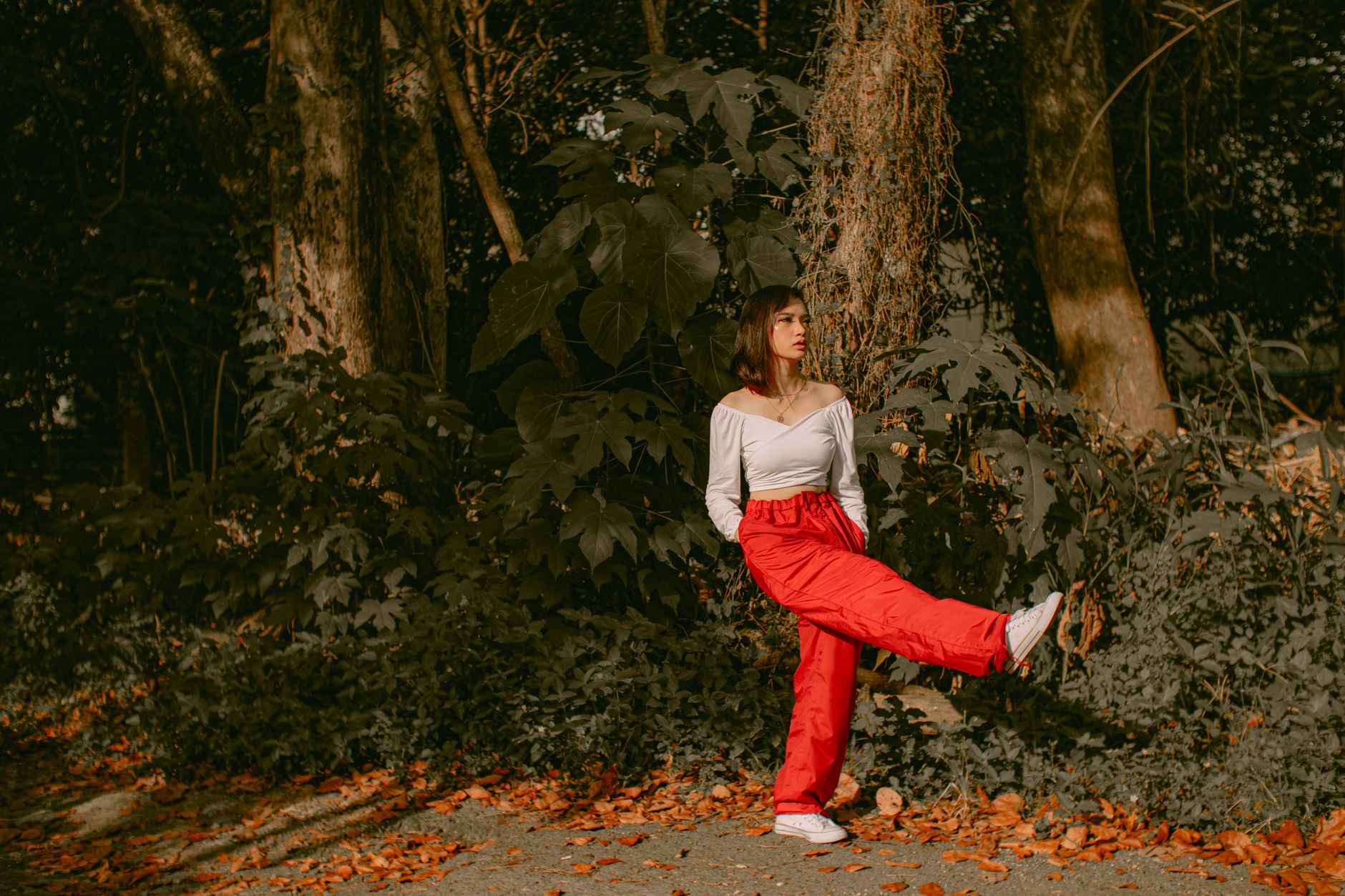 Full body of young female in trendy clothes standing with hands in pockets and raising leg in park with autumn foliage in sunny day