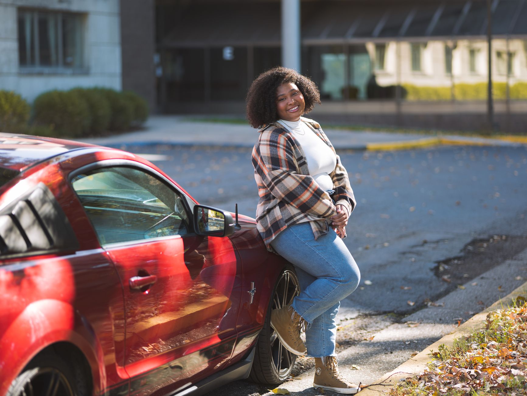 Plus-size African American woman with curly hair leaning against a red car in a relaxed outdoor setting.