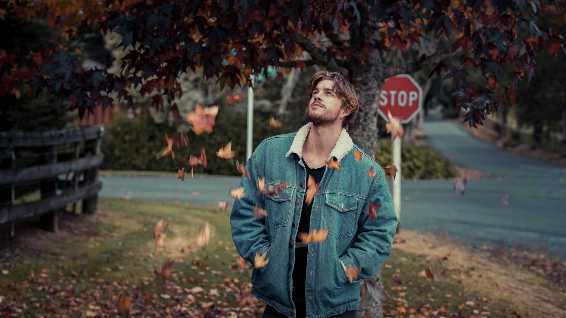 A man in a denim jacket enjoying falling autumn leaves on a roadside.