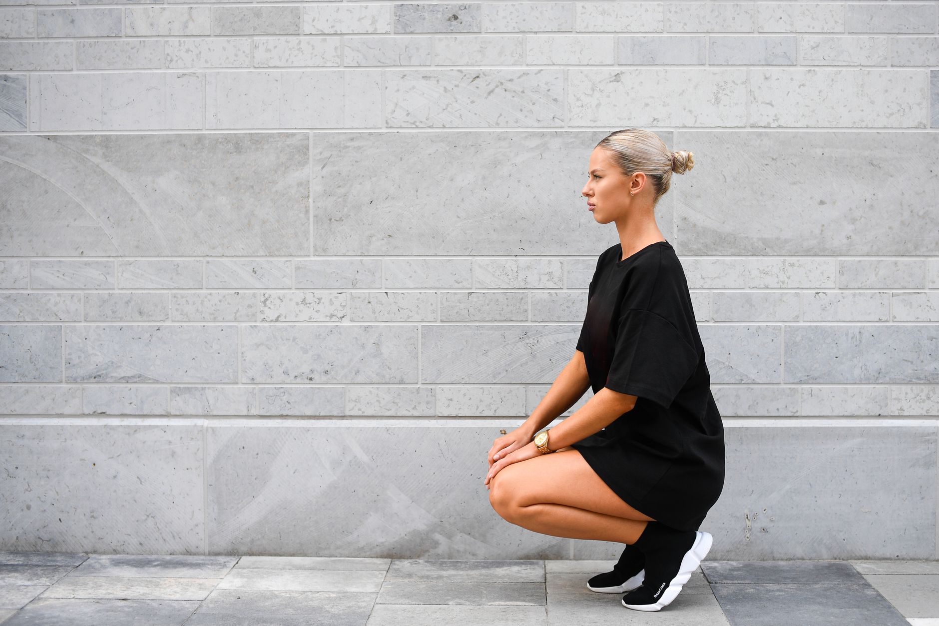 A fashionable young woman squats in modern streetwear against a minimalist stone wall in Tallinn, Estonia.