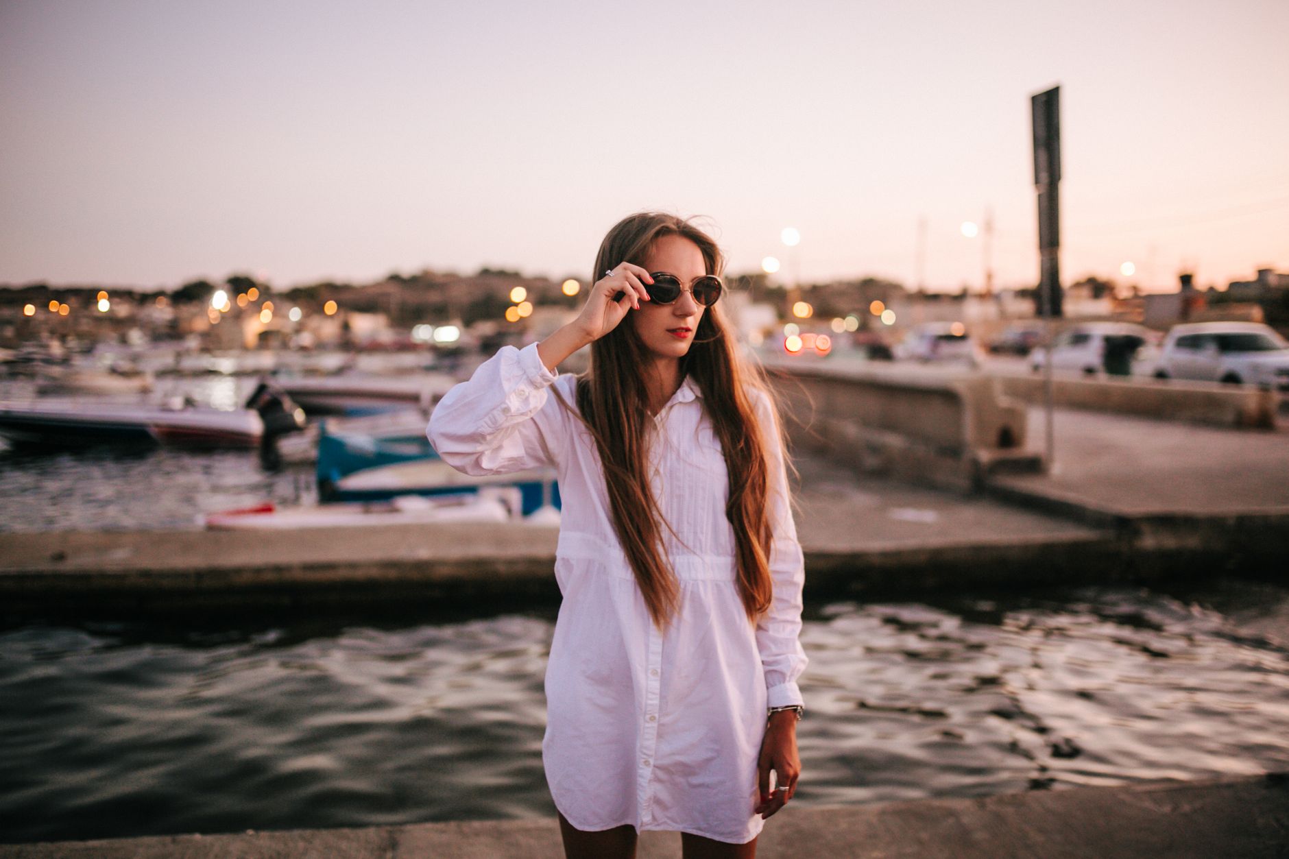 Fashionable woman in sunglasses standing by a harbor during sunset, creating a serene and stylish scene.