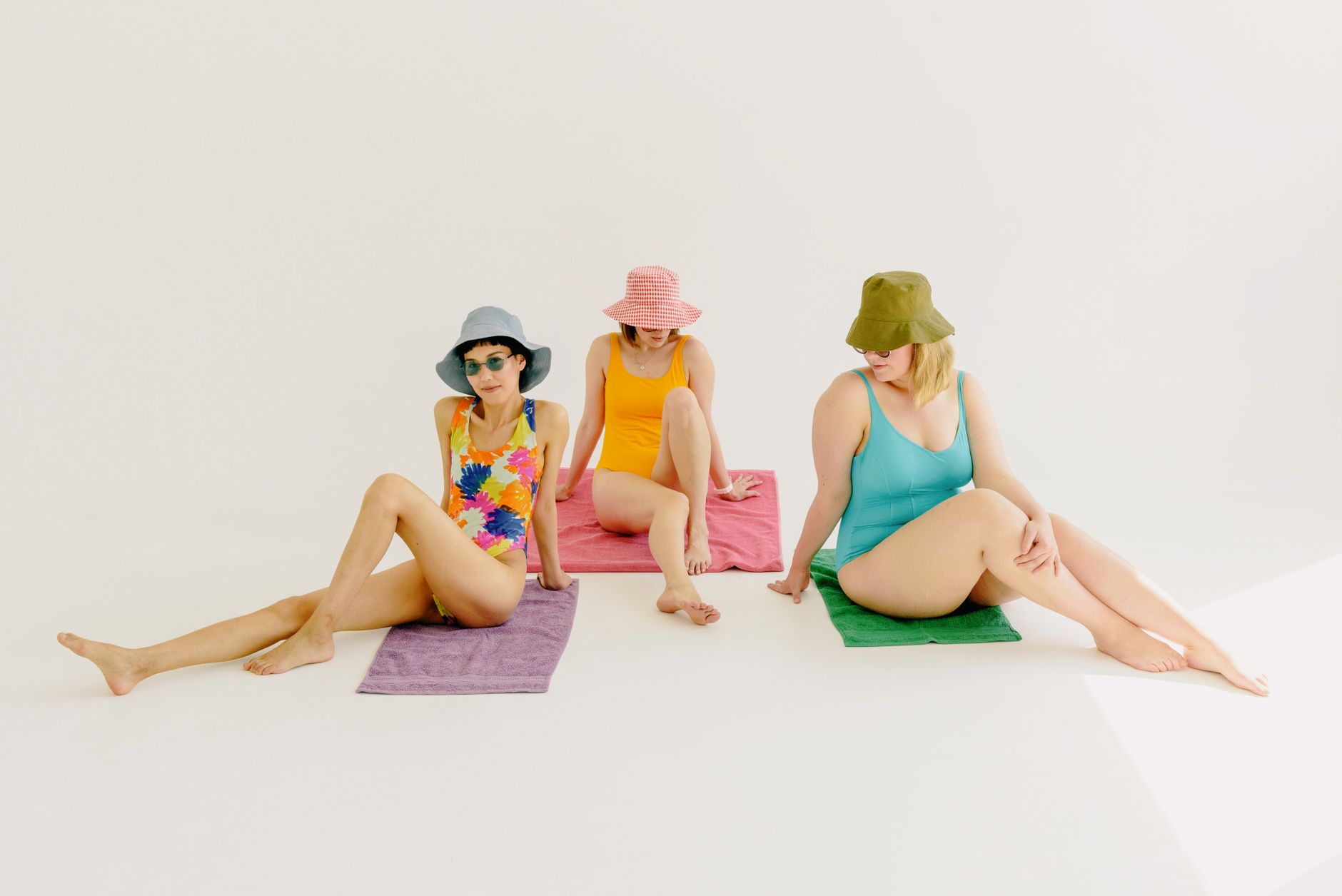 Three women in vibrant swimsuits and bucket hats sit on towels in a bright, minimalist studio setup.