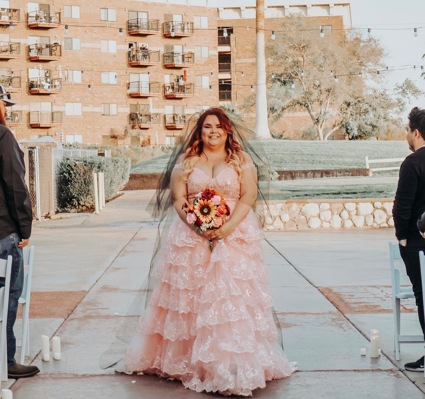 A smiling bride in a pink wedding dress poses outdoors in an urban setting.