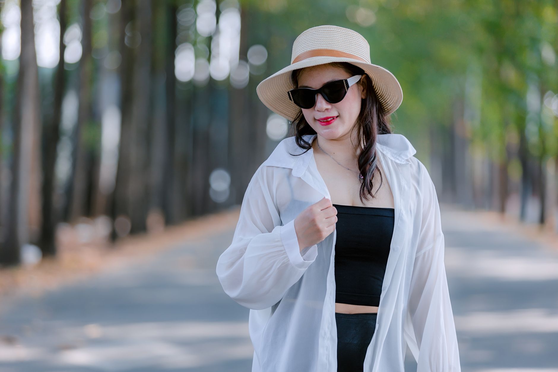 Fashionable woman wearing a sunhat and sunglasses walks confidently on a tree-lined path.