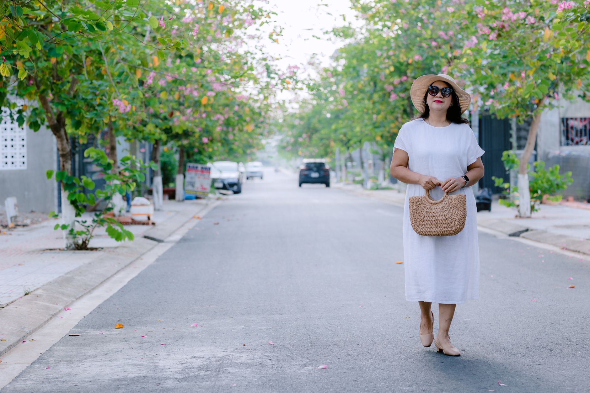 Stylish woman in white dress walking on a tree-lined street with blooming flowers.