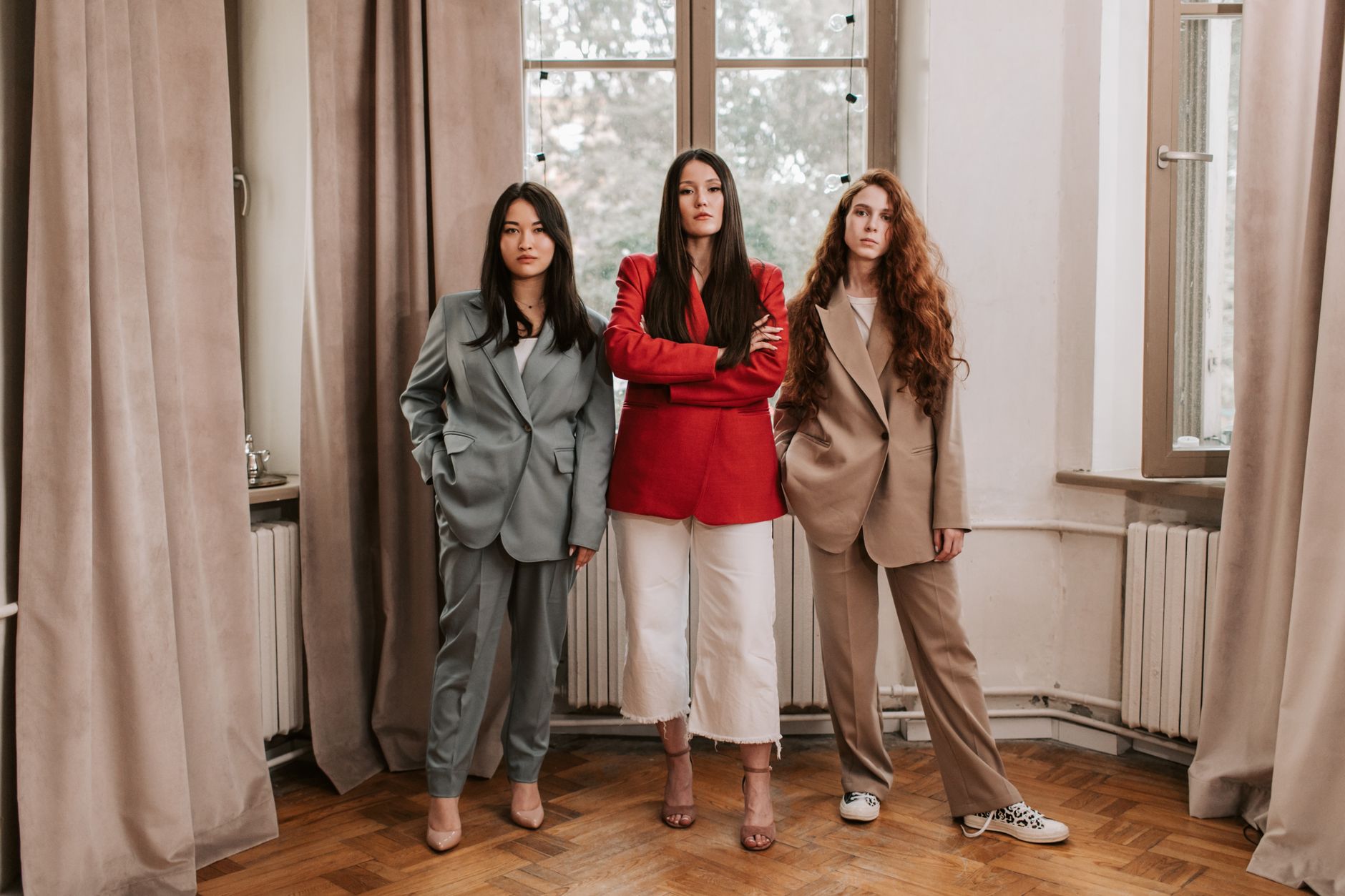 Three women confidently pose indoors, showcasing stylish business attire against elegant curtains.