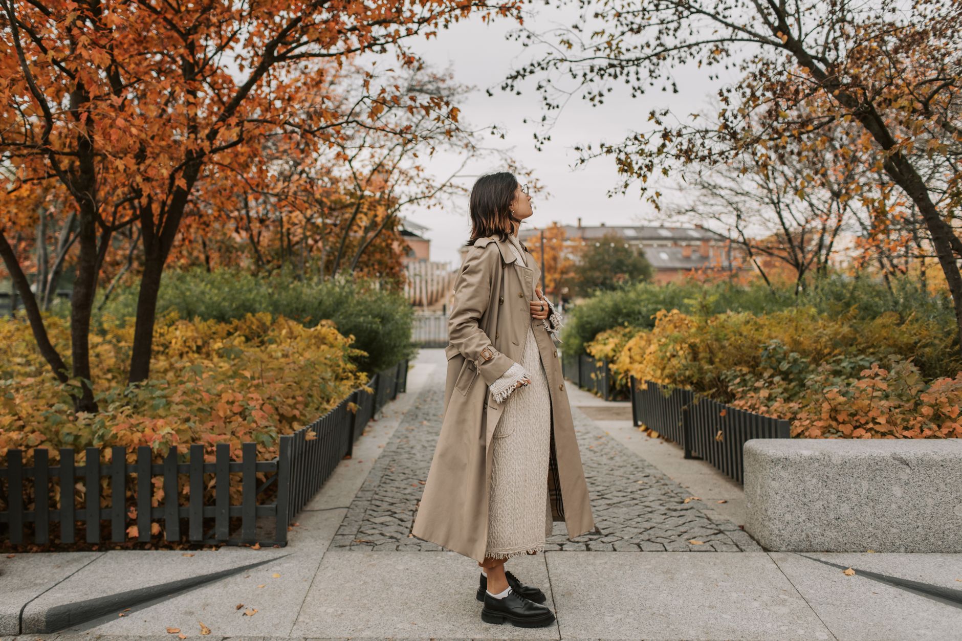 Woman in a trench coat walking through picturesque autumn parkway with vibrant foliage.