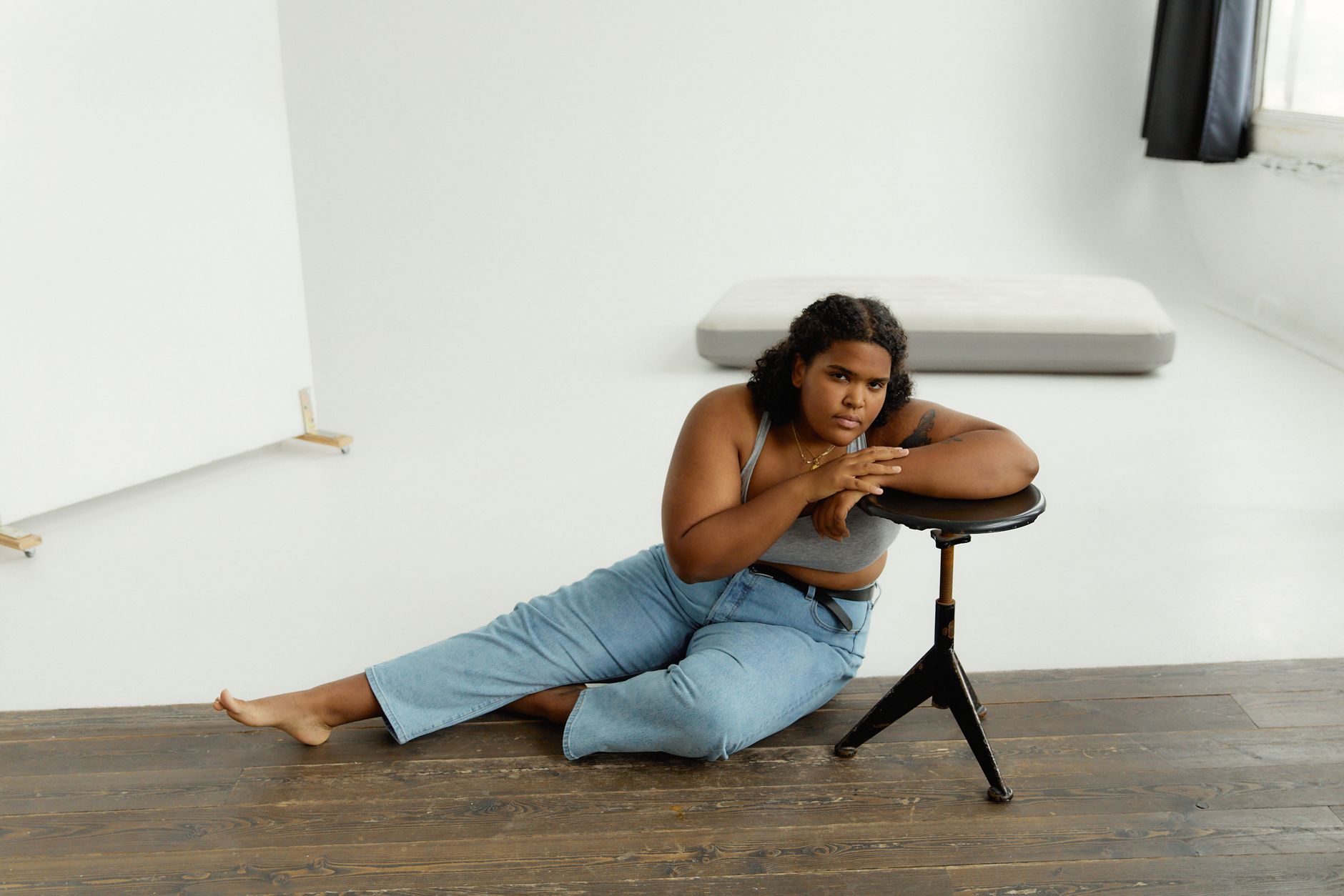 Plus size woman in denim jeans, bra top, leaning on a stool in a bright studio.