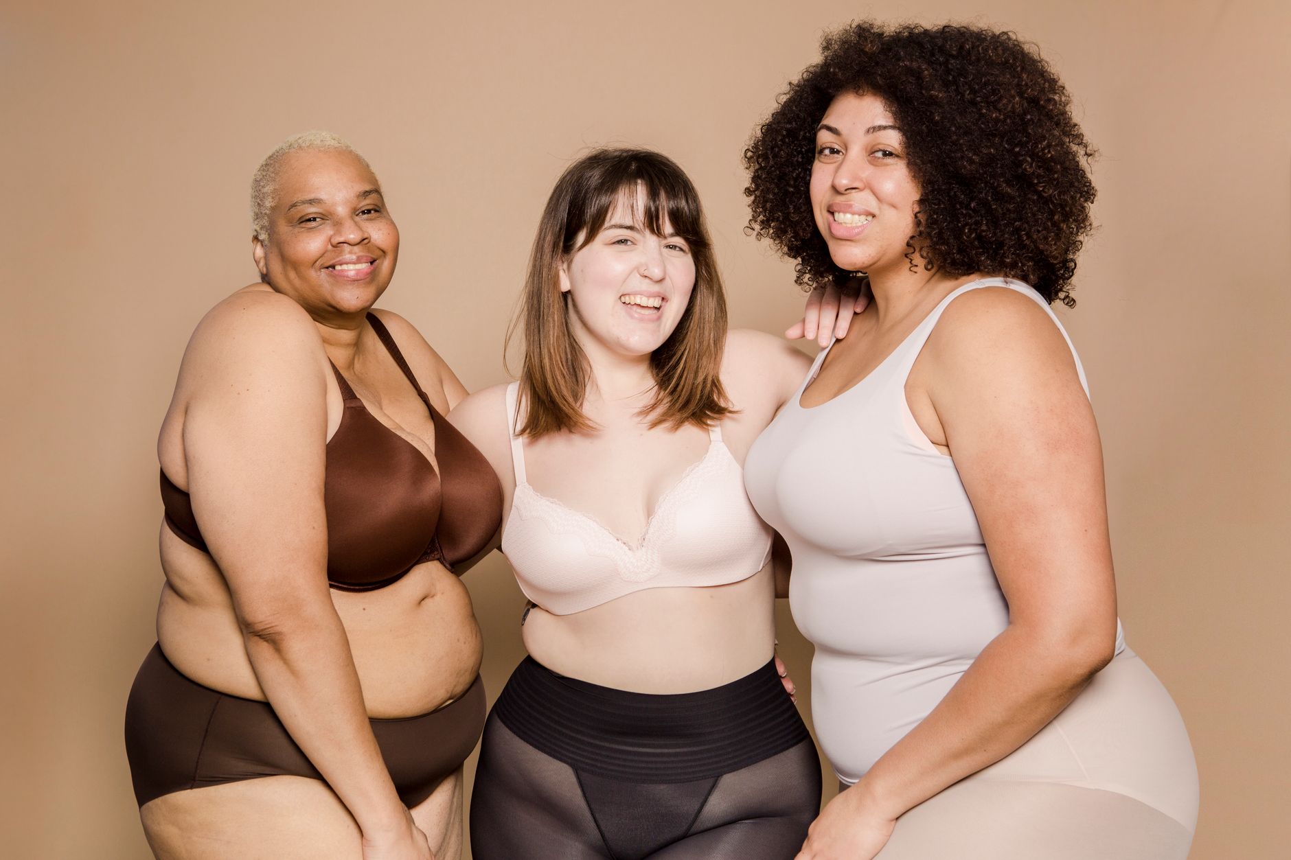 Group of cheerful young multiracial plus size female models in underwear smiling and looking at camera while cuddling against beige backgrou