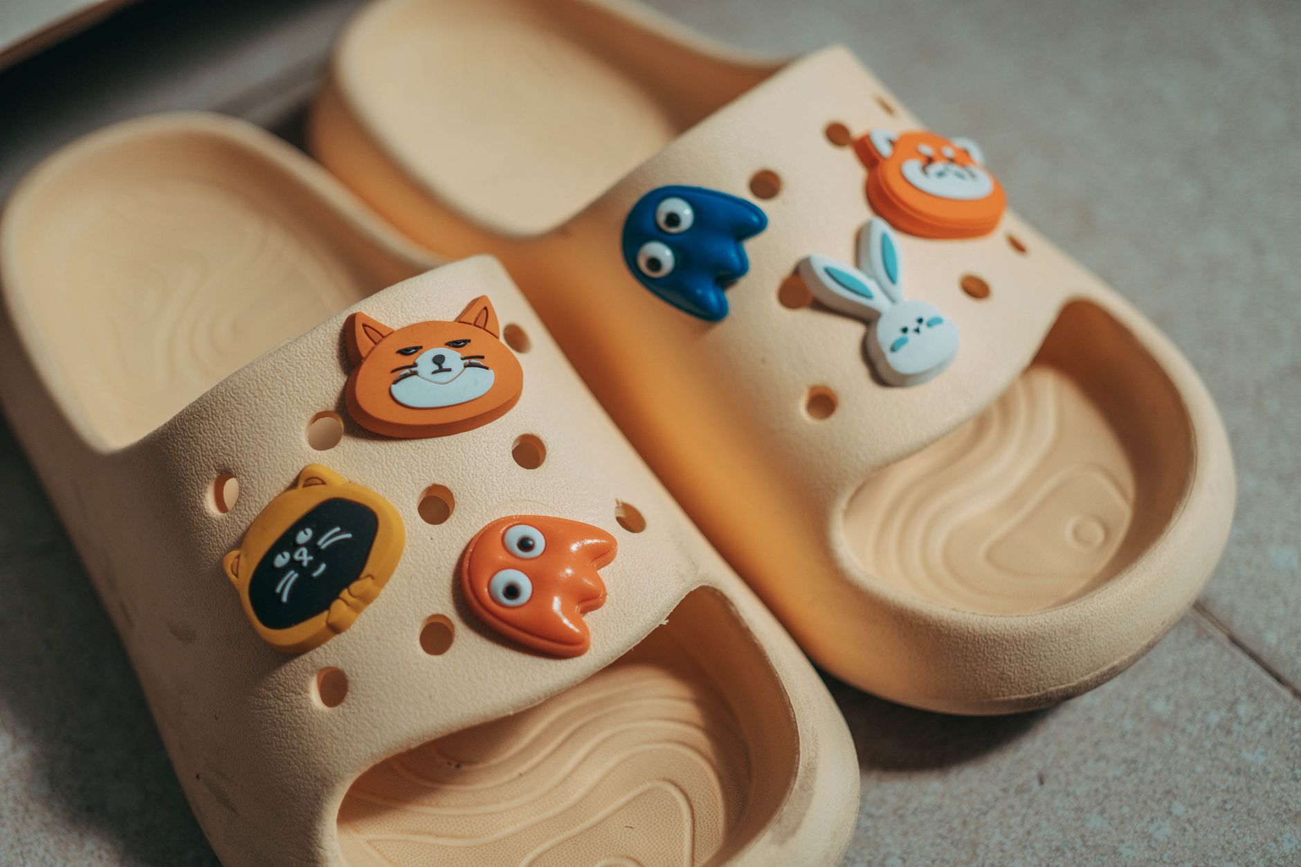 Beige croc sandals adorned with fun and colorful animal-shaped charms on a tiled floor.