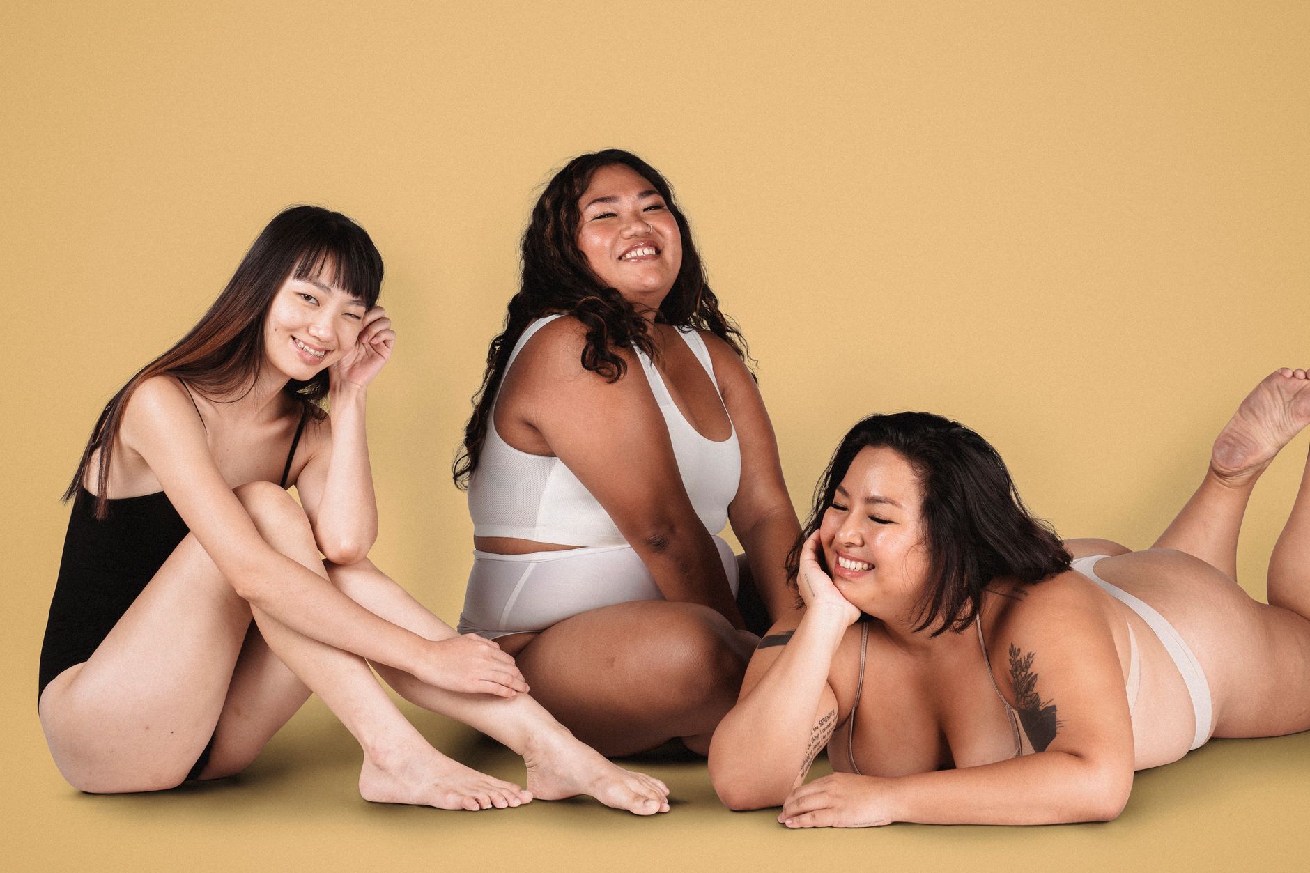 Cheerful Asian plus size models wearing white underwear while sitting near skinny smiling woman in black bodysuit on yellow background