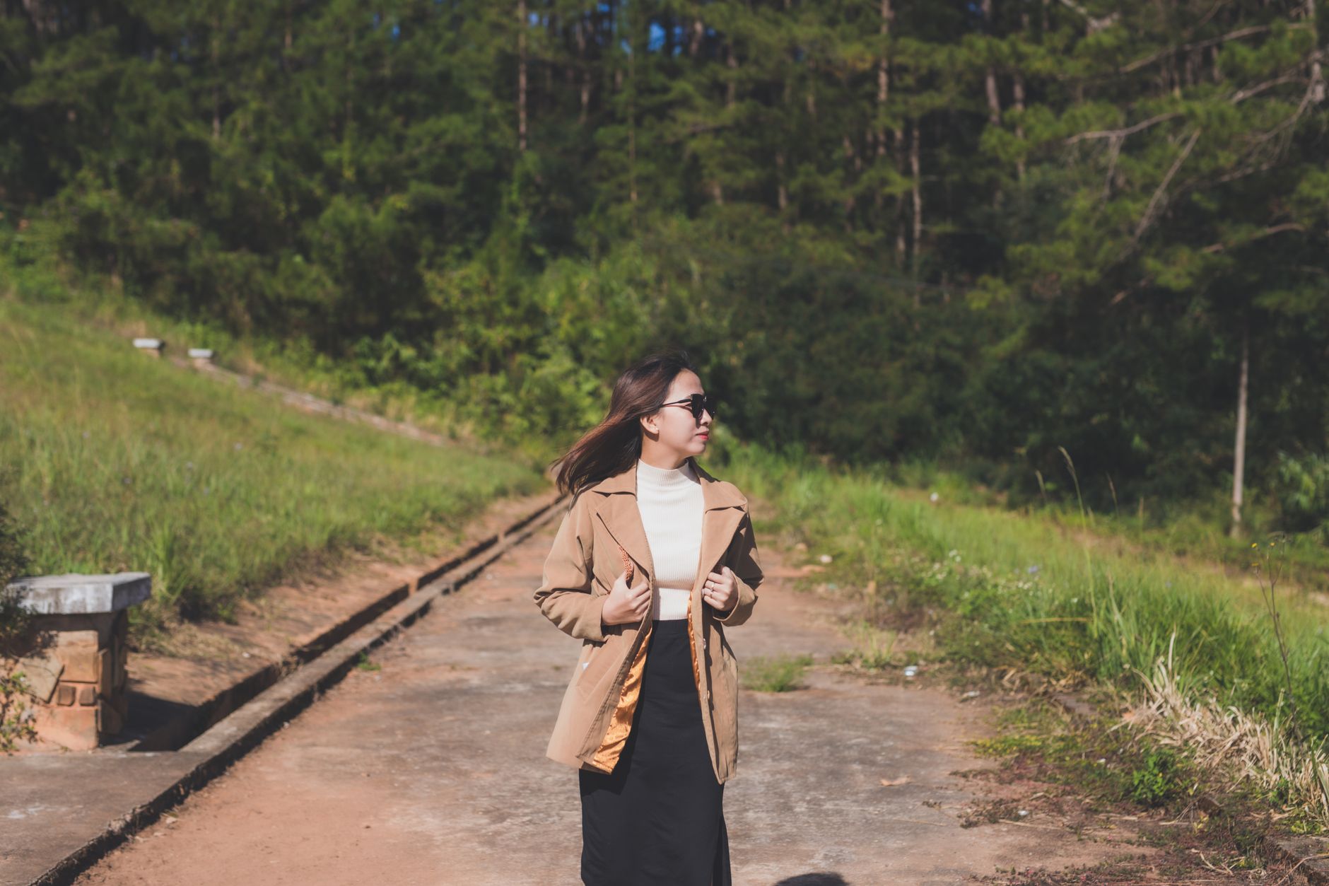 Fashionable woman in brown jacket strolling on a scenic path with sunglasses.