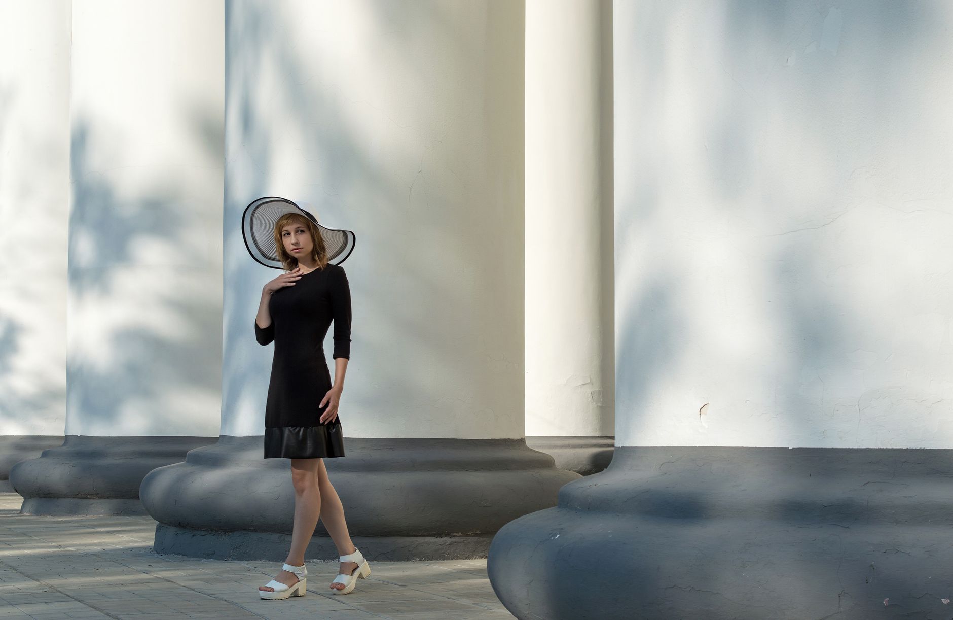 Sophisticated woman in black dress and large hat posing by classical columns outdoors.