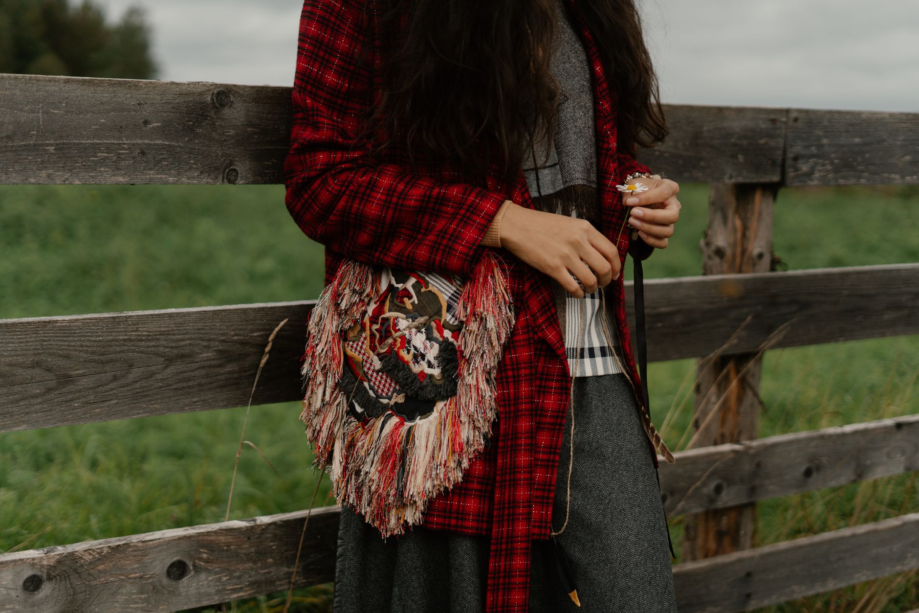 A woman in boho attire with an embroidered bag leaning on a wooden fence outdoors.