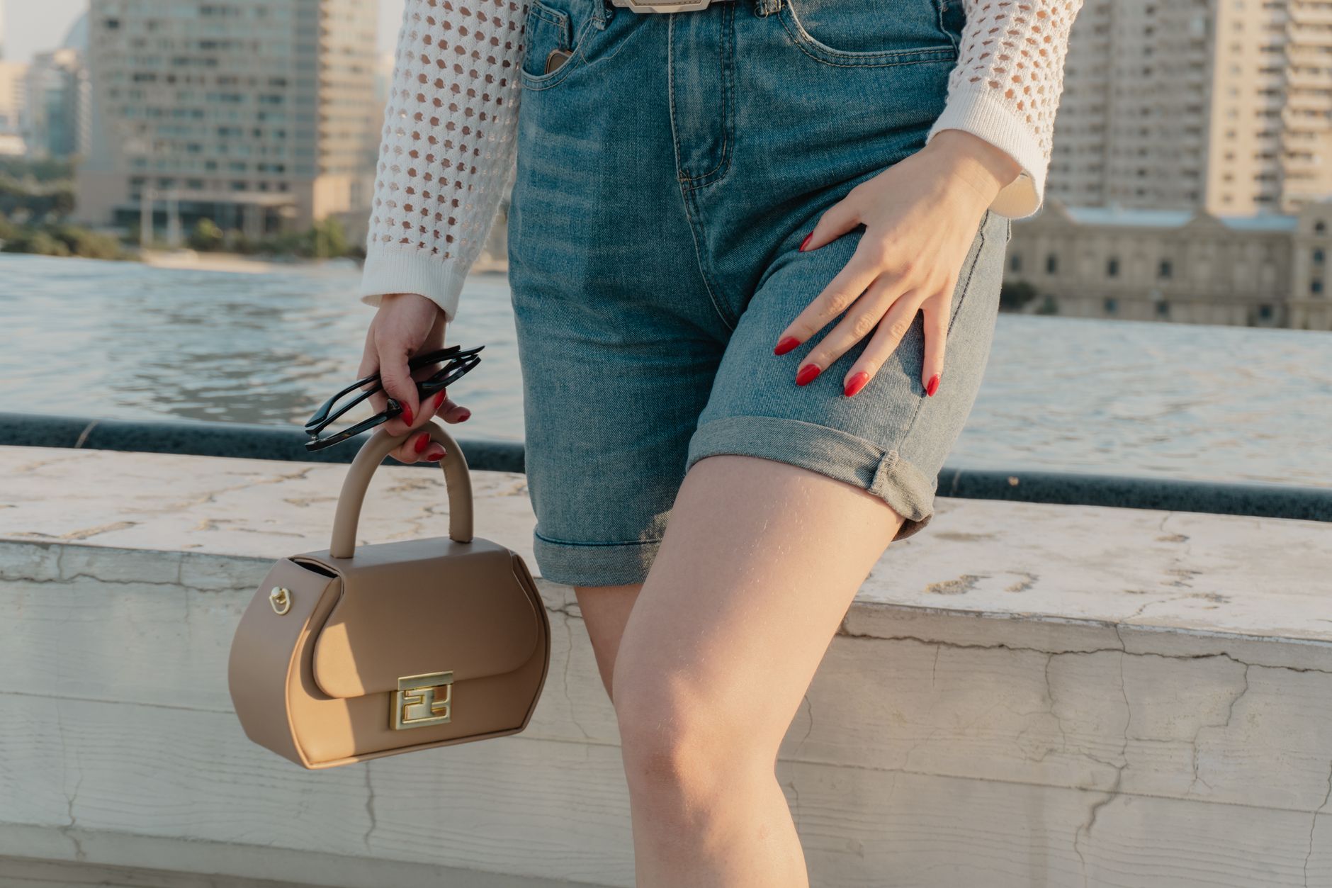 Stylish woman on rooftop wearing denim shorts, holding a handbag, showcasing red nails.