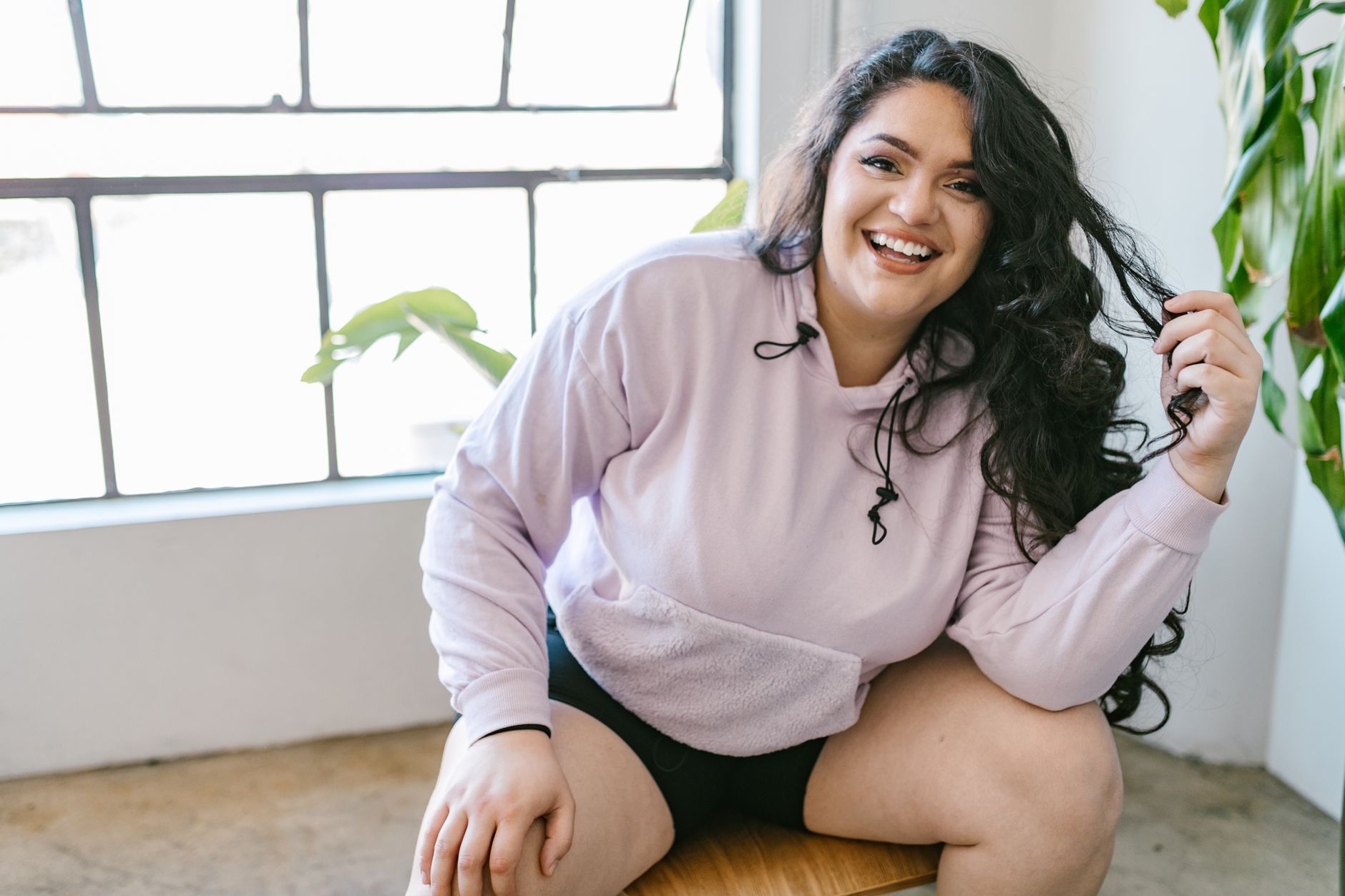 A confident plus-size woman in a stylish hoodie smiles indoors, embodying self-love and body positivity.