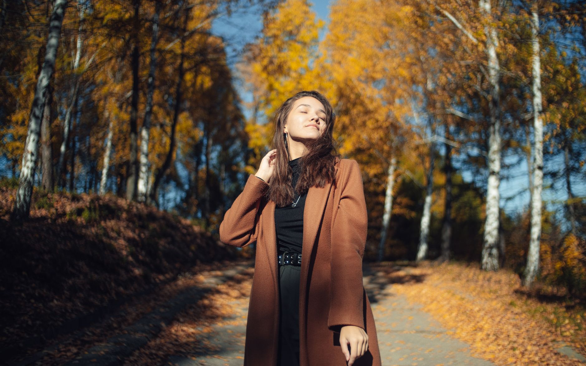 A woman in a brown coat enjoys a sunny autumn day in a birch forest.