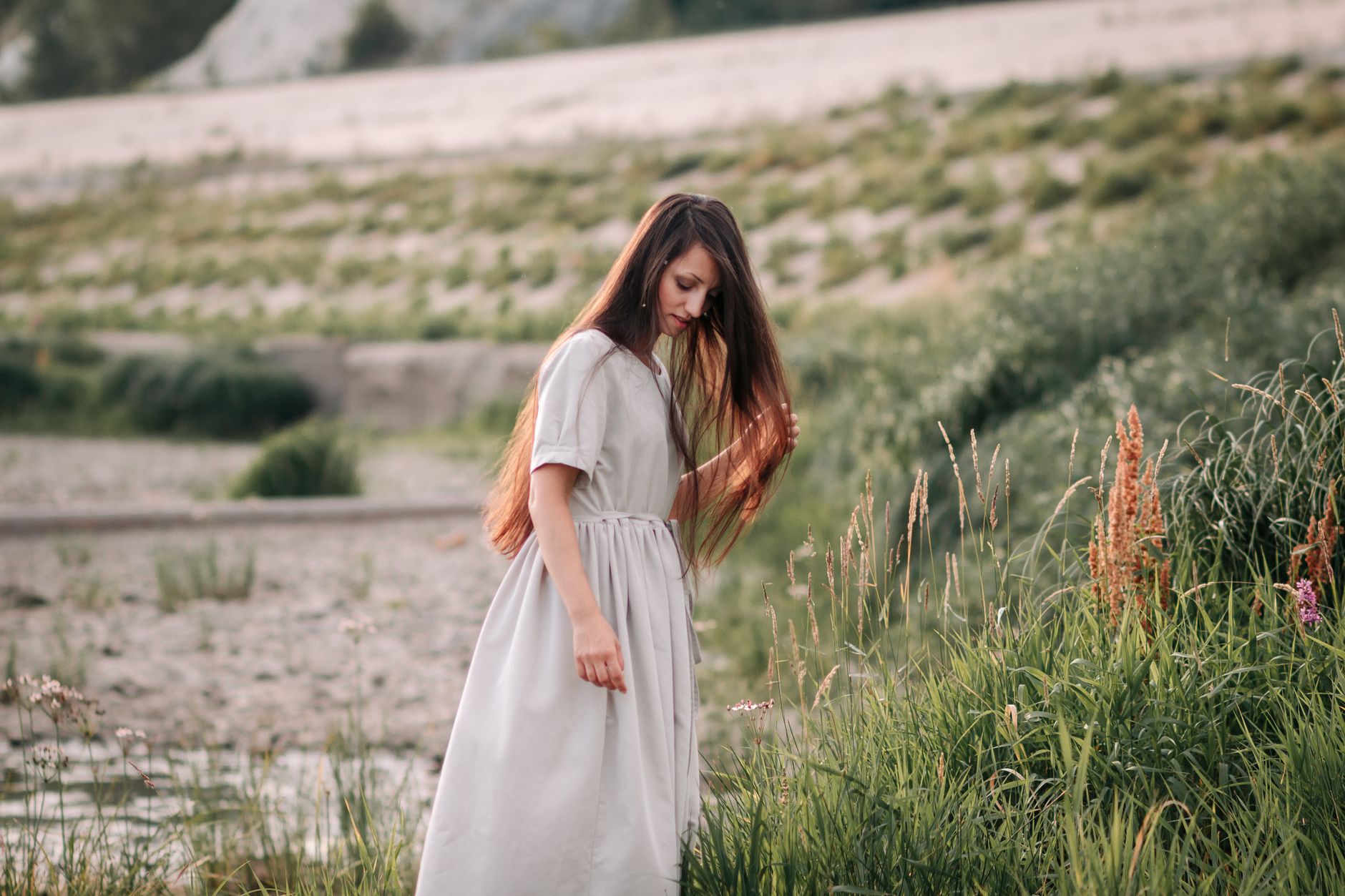 Peaceful scene of a woman in a flowing beige dress walking thoughtfully through a lush countryside setting.