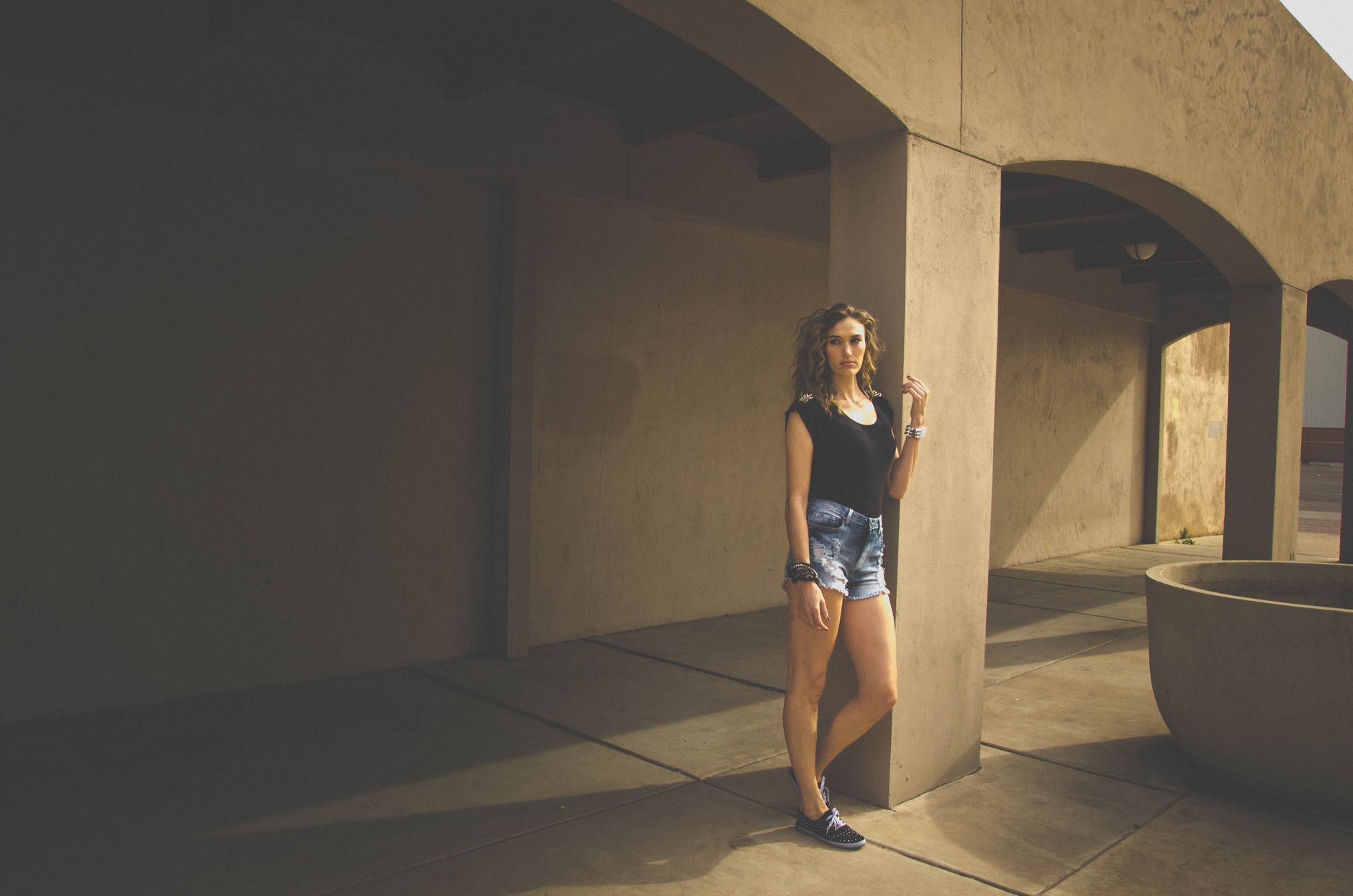 Stylish young woman in denim shorts poses outdoors in an urban architectural setting.