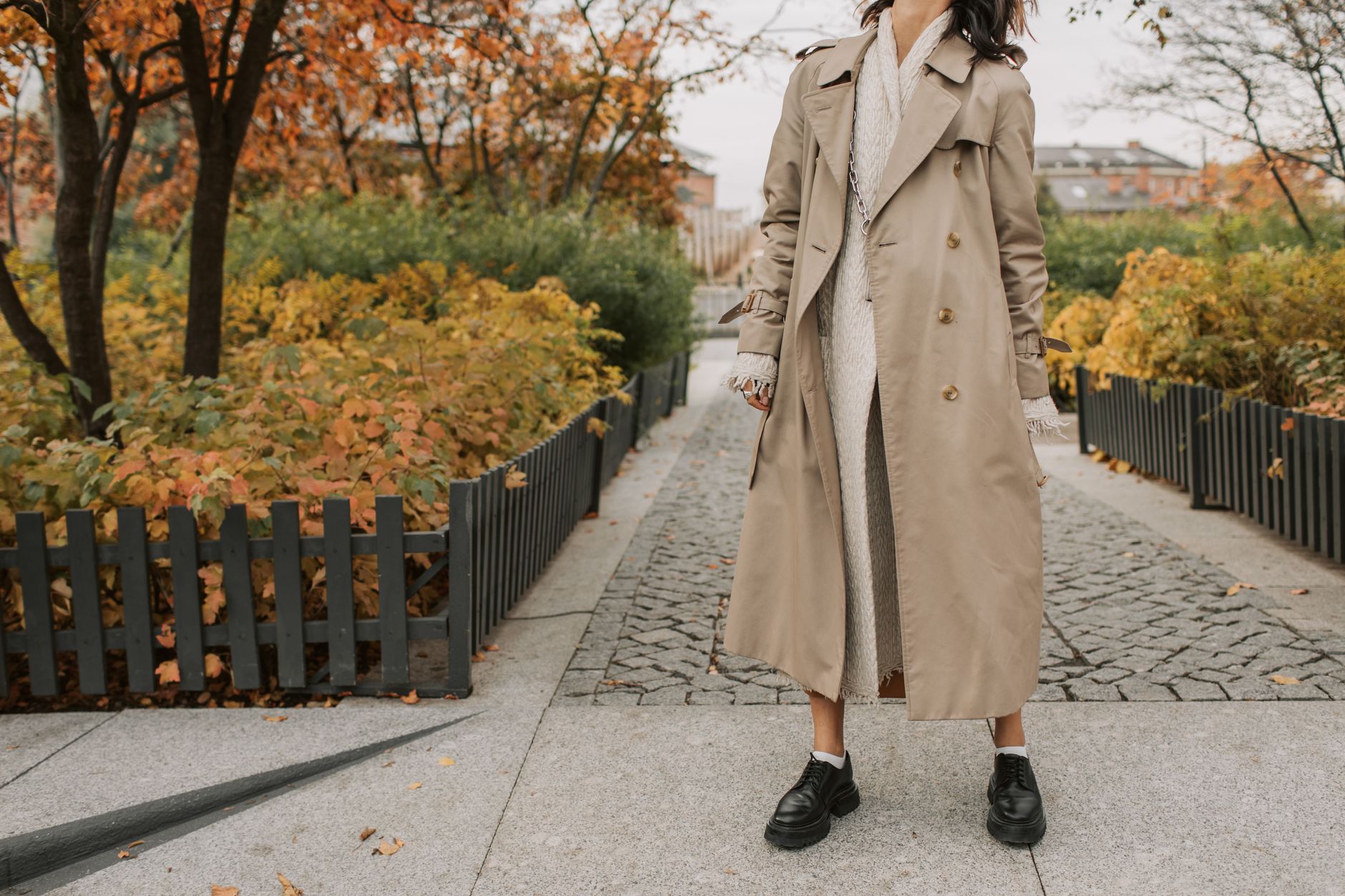 A fashionable woman stands in a park wearing a trench coat, surrounded by autumn foliage.