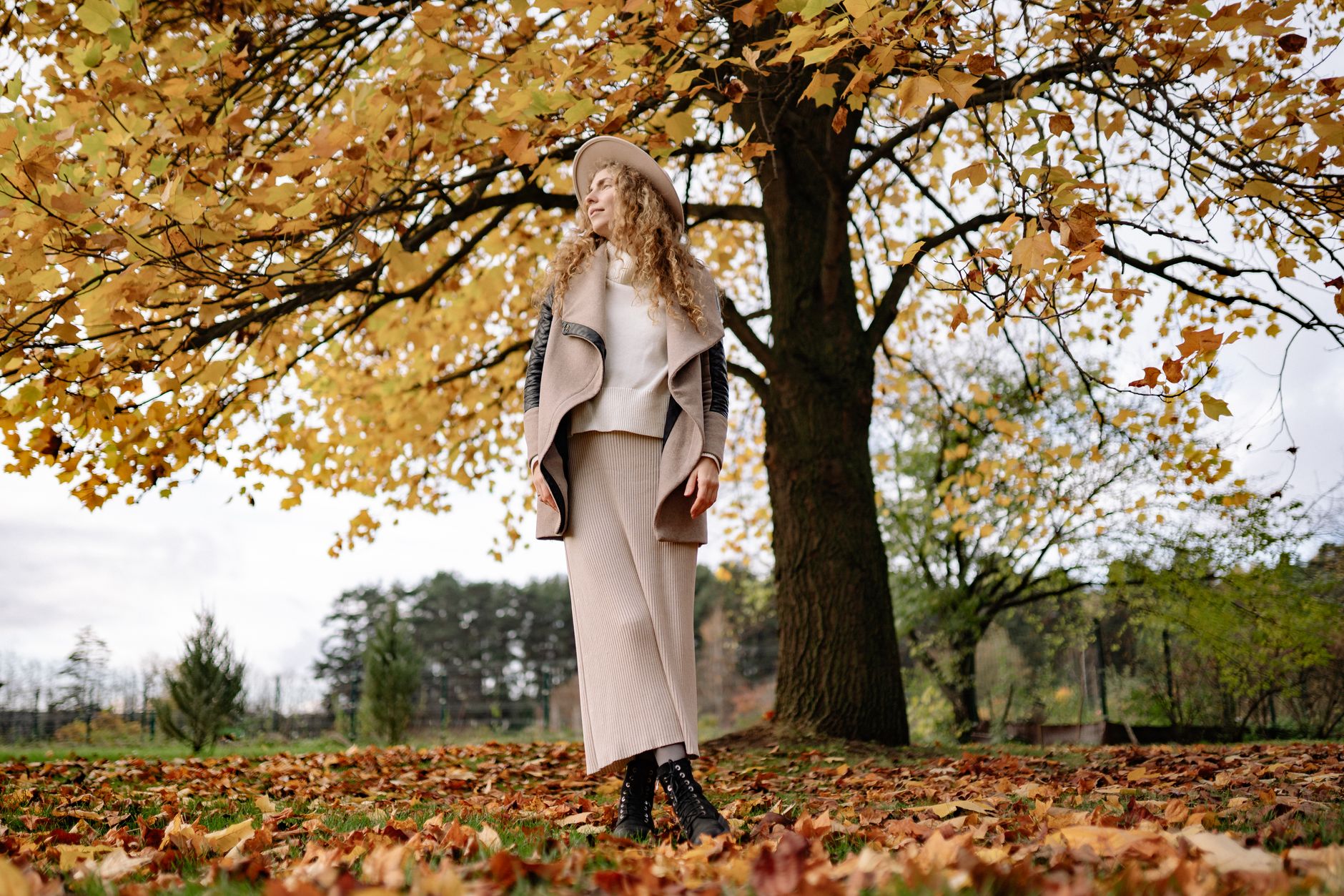 Woman posing under autumn leaves in stylish attire, showcasing fall fashion.
