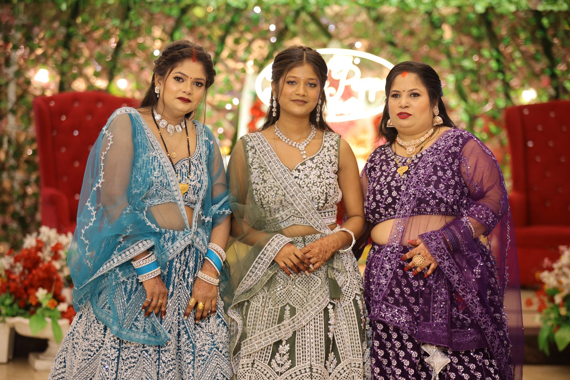 Three women in beautiful traditional outfits posing at a decorated indoor event.