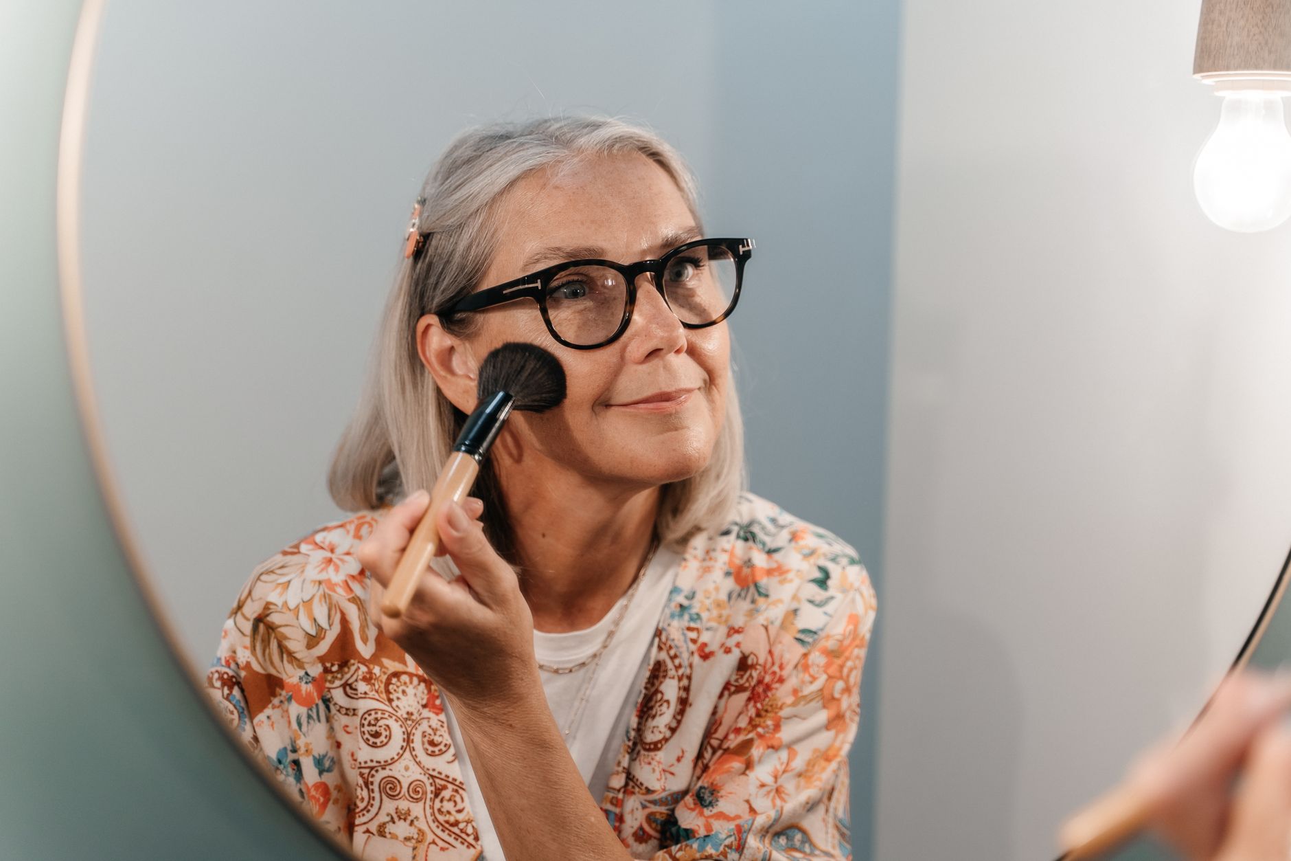 Senior woman with glasses applying makeup while looking in a mirror, reflecting a joyful moment.
