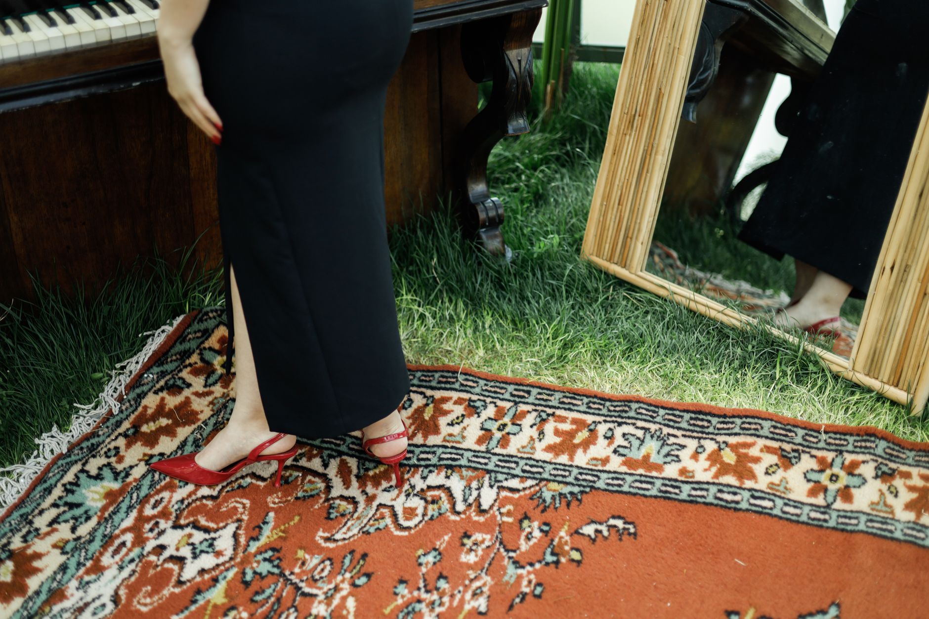 Elegant woman in black dress and red heels beside piano and mirror on patterned carpet.