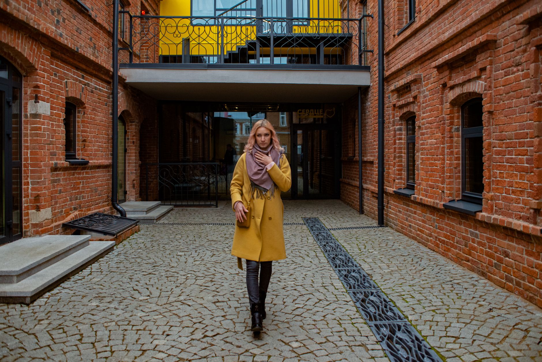 Stylish woman in yellow coat and scarf strolling through a modern brick alleyway.