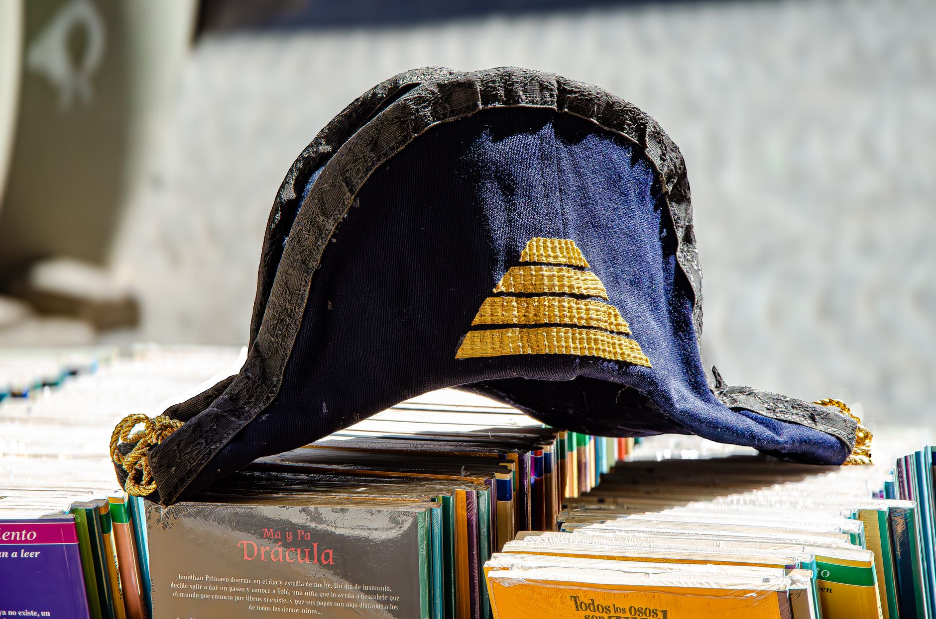 A vintage bicorn hat with golden details resting on a stack of books outdoors.