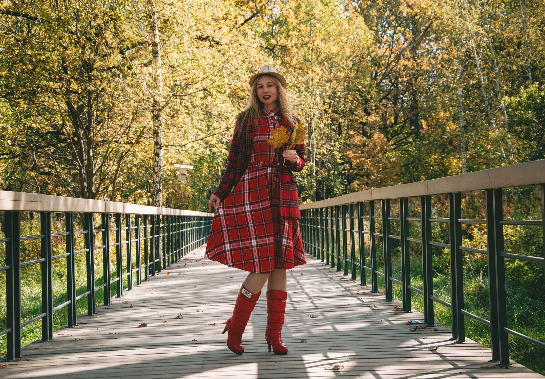 Charming woman in red plaid dress and boots walking on a scenic wooden bridge amidst colorful autumn foliage.