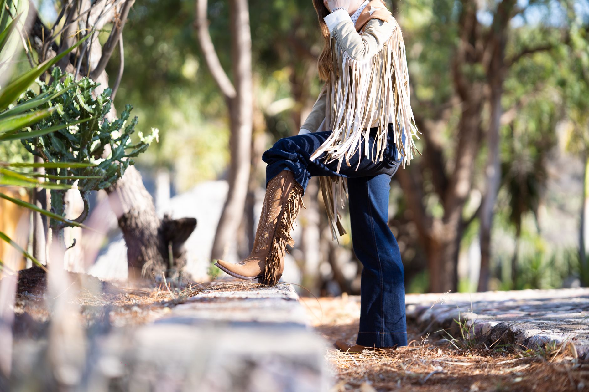 A stylish woman with a fringe jacket poses outdoors amidst trees during fall.