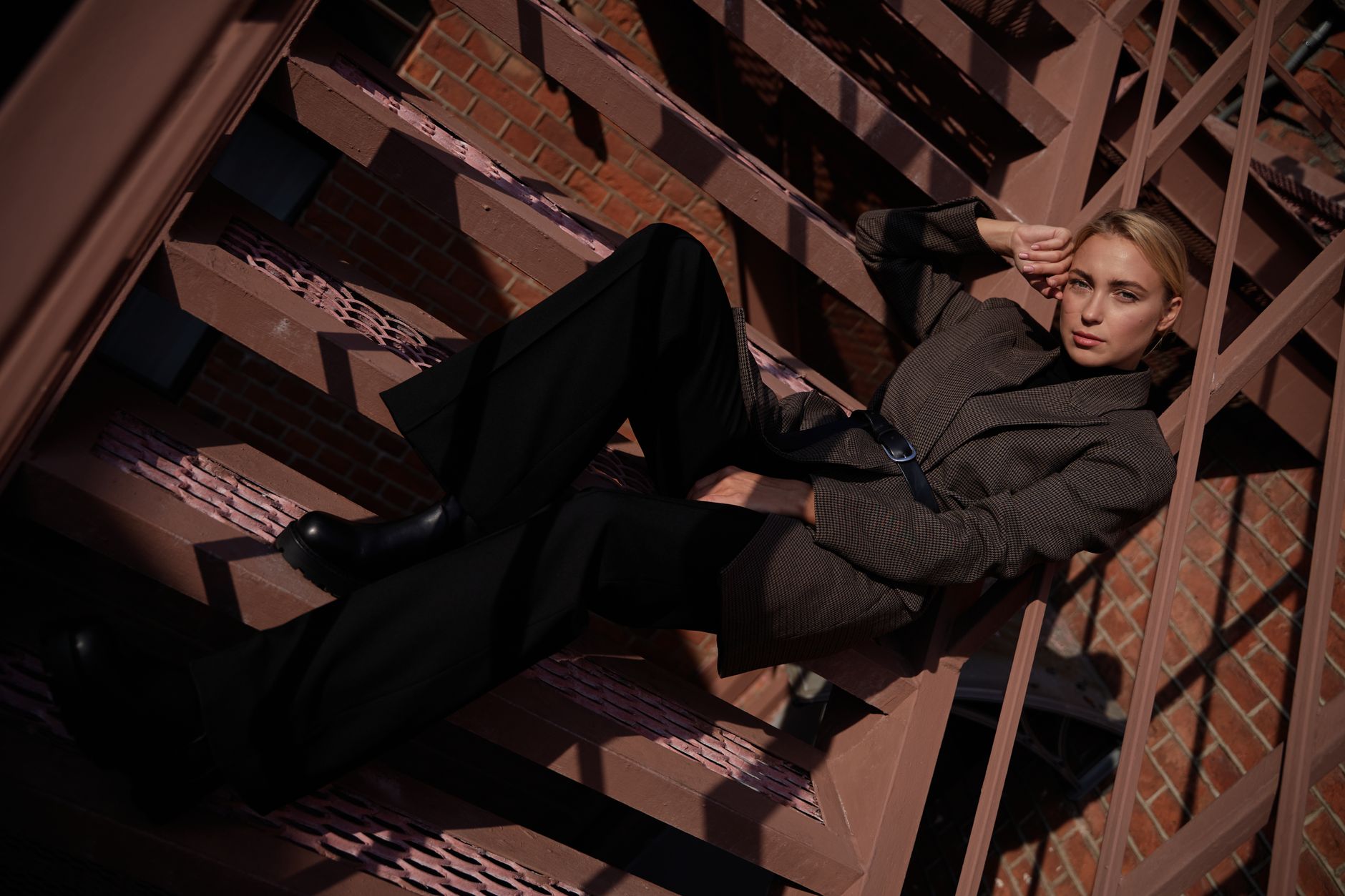 Stylish woman in a brown blazer lounging on fire escape stairs with urban background.