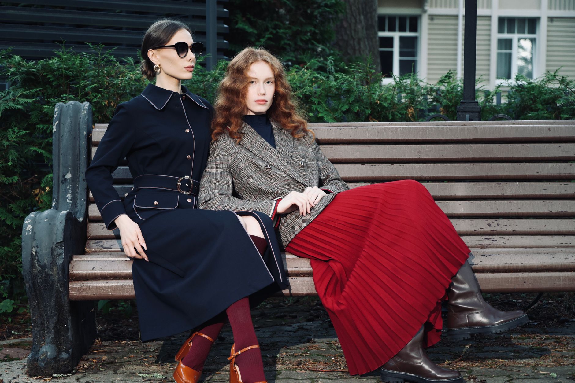 Two women in stylish fall outfits seated on a park bench, exuding elegance and sophistication.