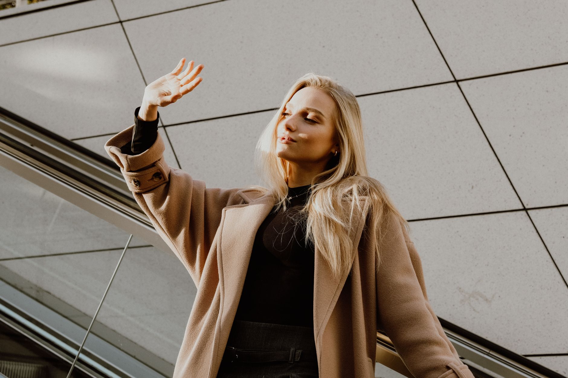 A blonde woman elegantly poses in a wool winter coat, enjoying a sunny day outdoors.