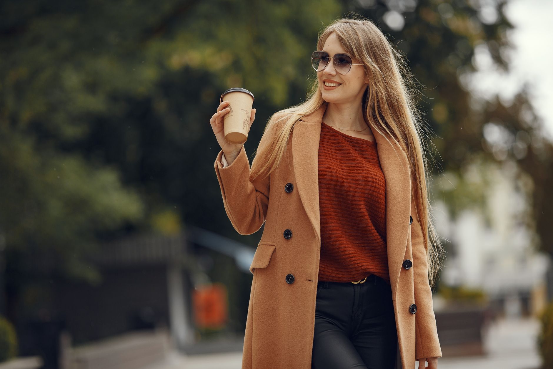 Stylish woman in a camel coat enjoying coffee on a sunny day outdoors.