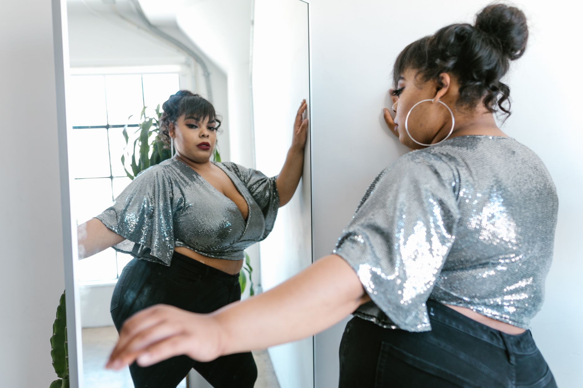 Elegant plus size model in a silver top posing confidently in front of a mirror indoors.